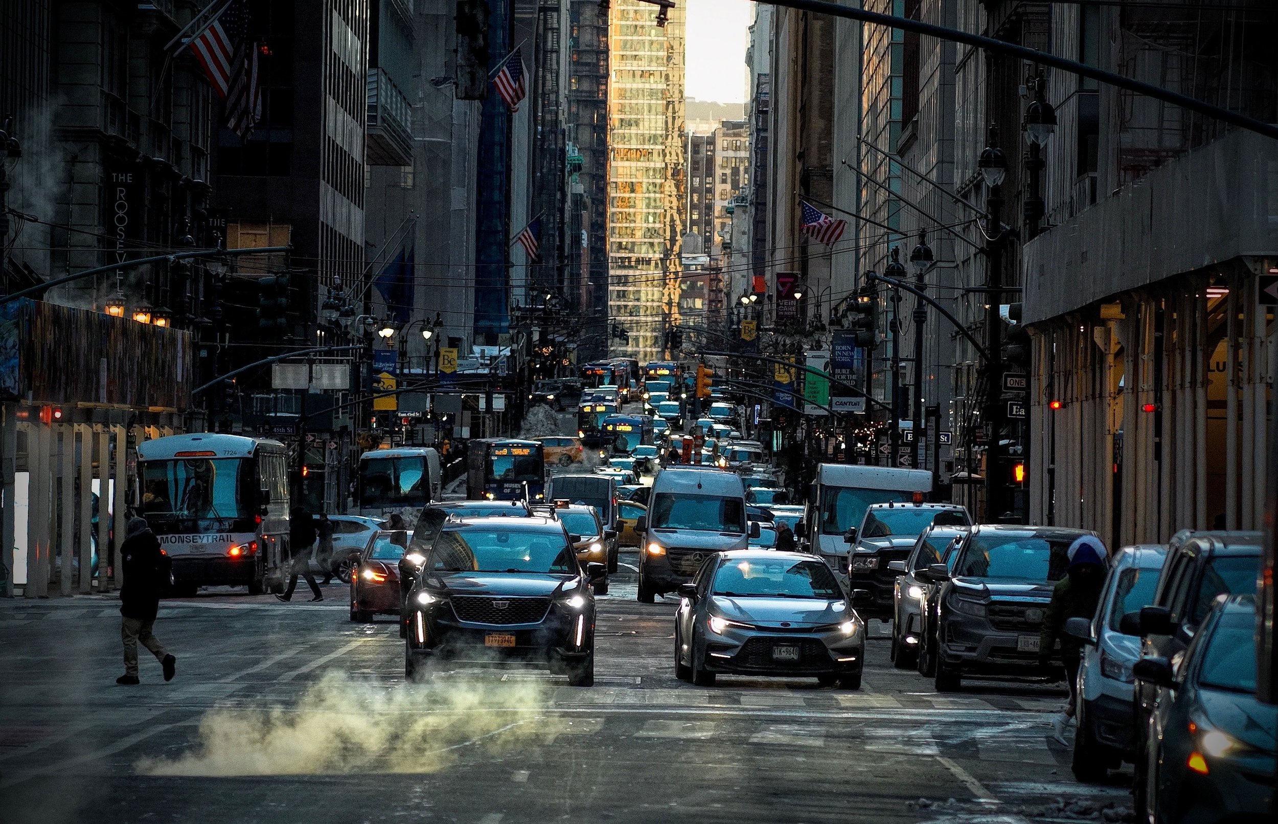 A busy city street in New York City filled with cars, buses, and pedestrians. Tall buildings line both sides of the street, with American flags hanging from some of the buildings. Steam rises from the street in the foreground, and the traffic extends