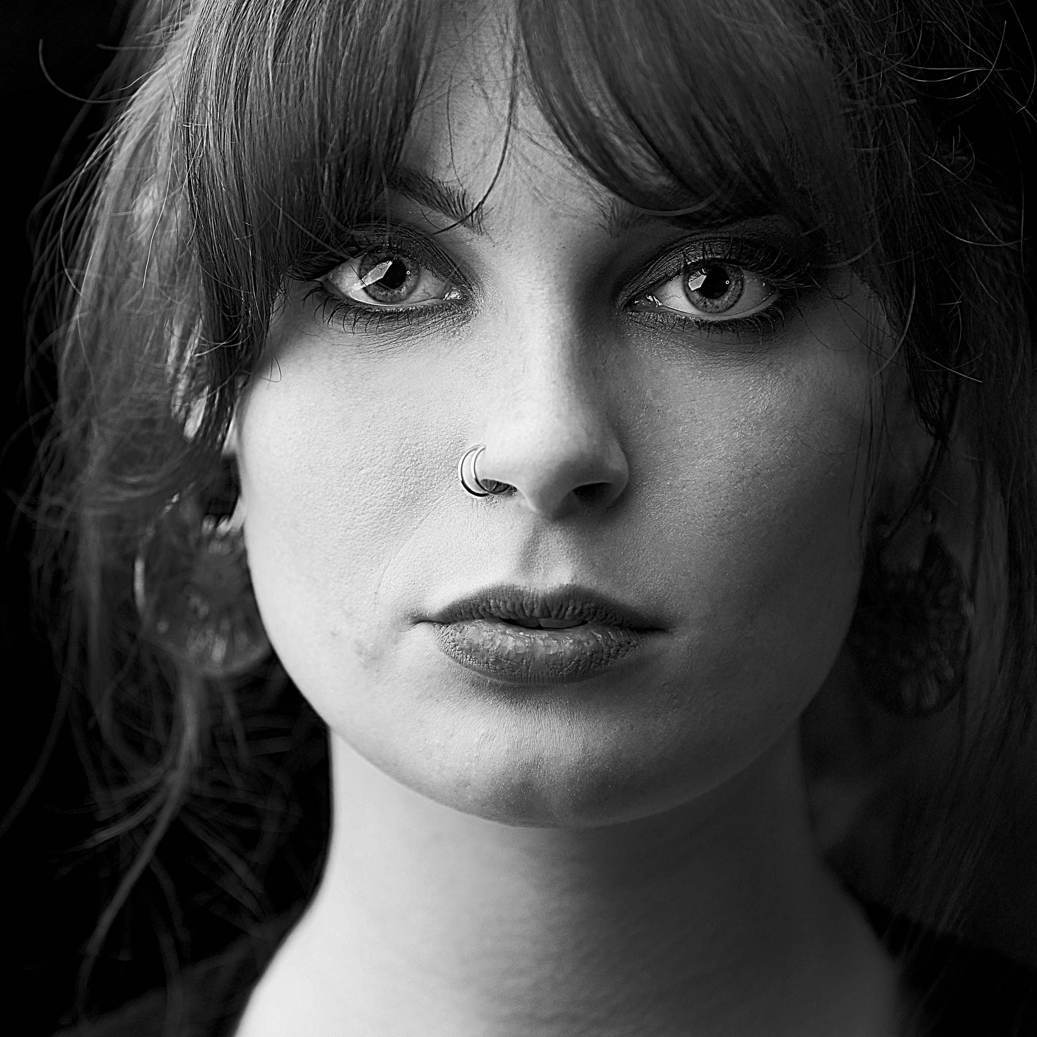Close-up black-and-white portrait of a woman with large eyes, dark hair, a nose ring, and full lips, gazing directly at the camera.