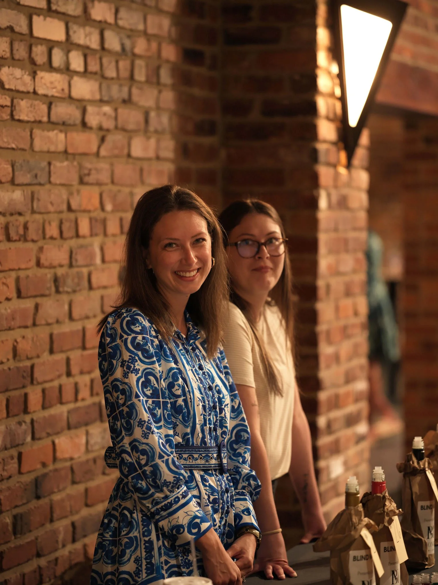 Two women standing behind a counter with bottles wrapped in brown paper bags. One woman wears a blue and white patterned dress, and the other wears glasses and a beige top. They are indoors against a brick wall, with warm lighting.