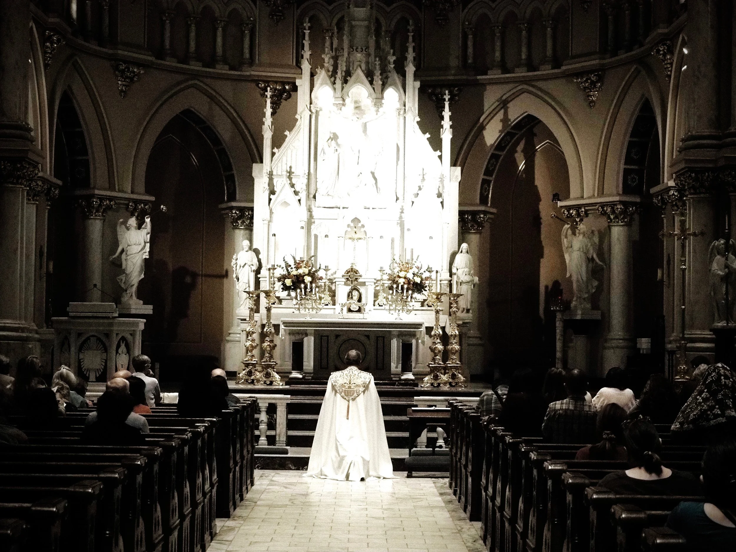 People seated in a church during a religious service or wedding ceremony, with an altar featuring statues, flowers, and ornate decorations in the background.