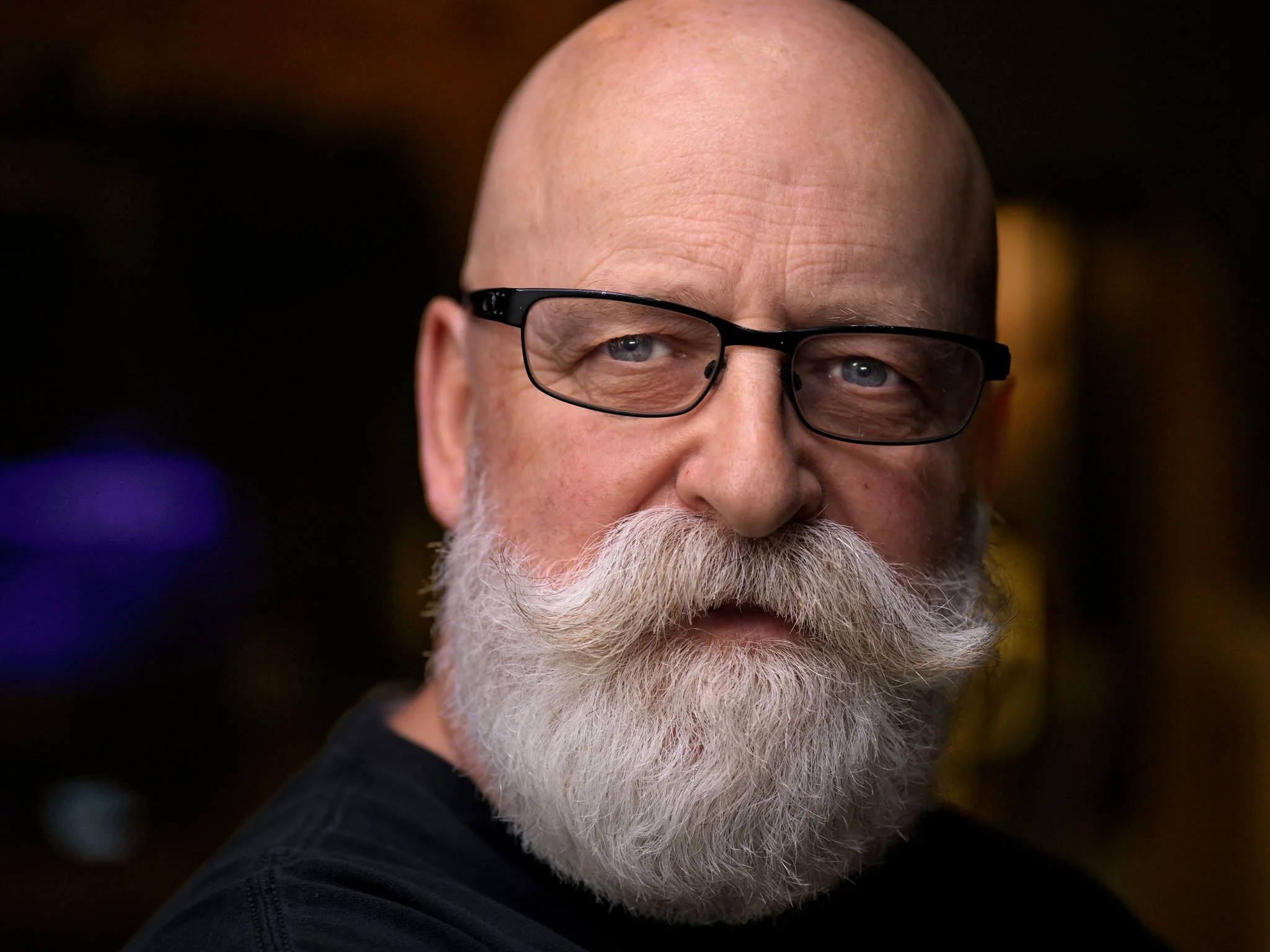 Close-up of an older man with a white beard, glasses, and a bald head, looking at the camera with a slight smile.