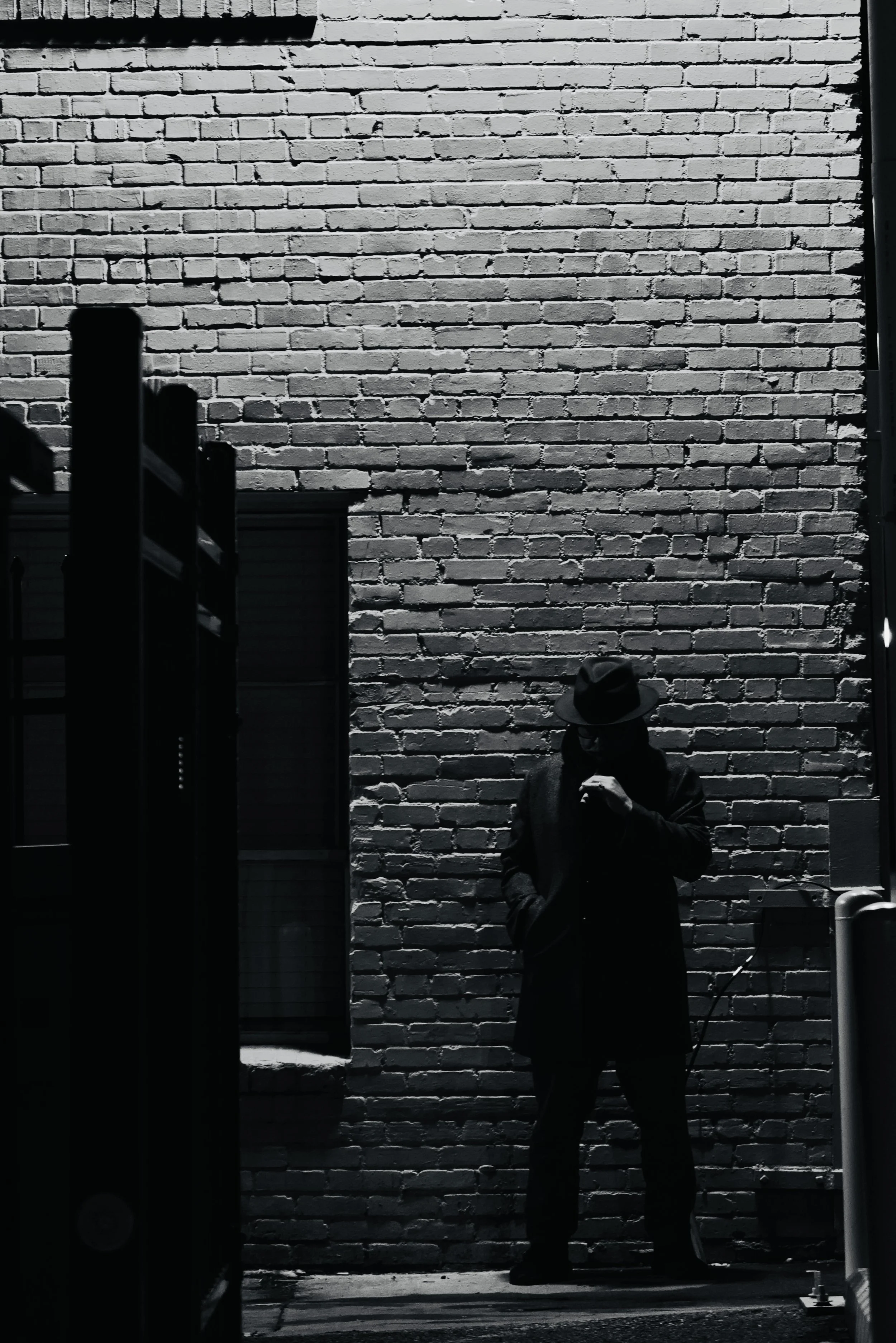 A person dressed in dark clothing and a fedora hat standing against a brick wall at night, holding a cigarette.