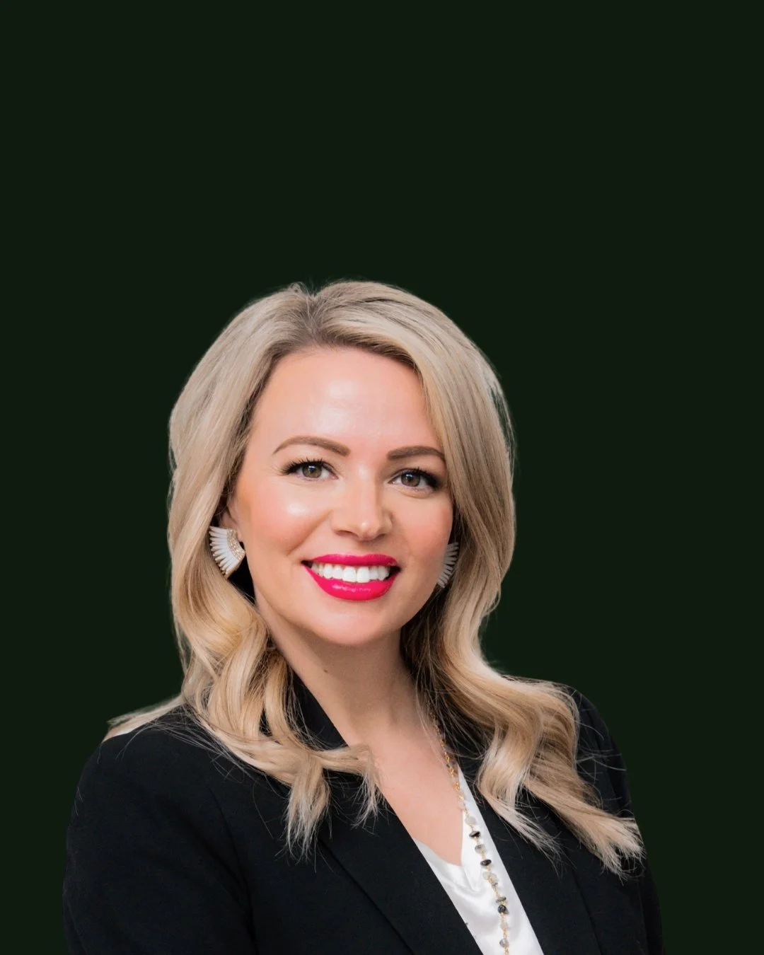 A woman with blonde hair styled in loose waves, wearing a black blazer, white top, statement earrings, and bright lipstick, smiling against a dark background.