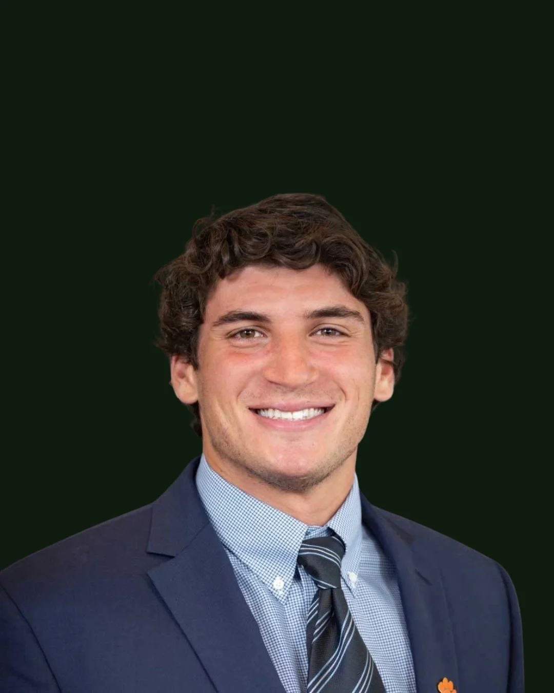 A young man with dark curly hair, smiling, wearing a dark blue suit, light blue checkered shirt, and striped tie, against a plain dark background.