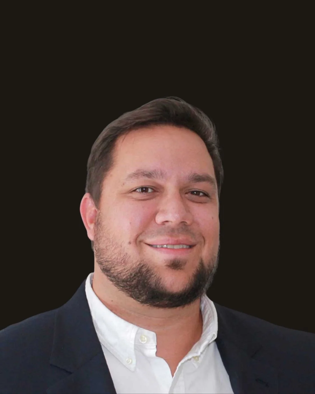 A man with brown hair, a beard, and a mustache, smiling, wearing a dark blazer and white shirt, against a black background.