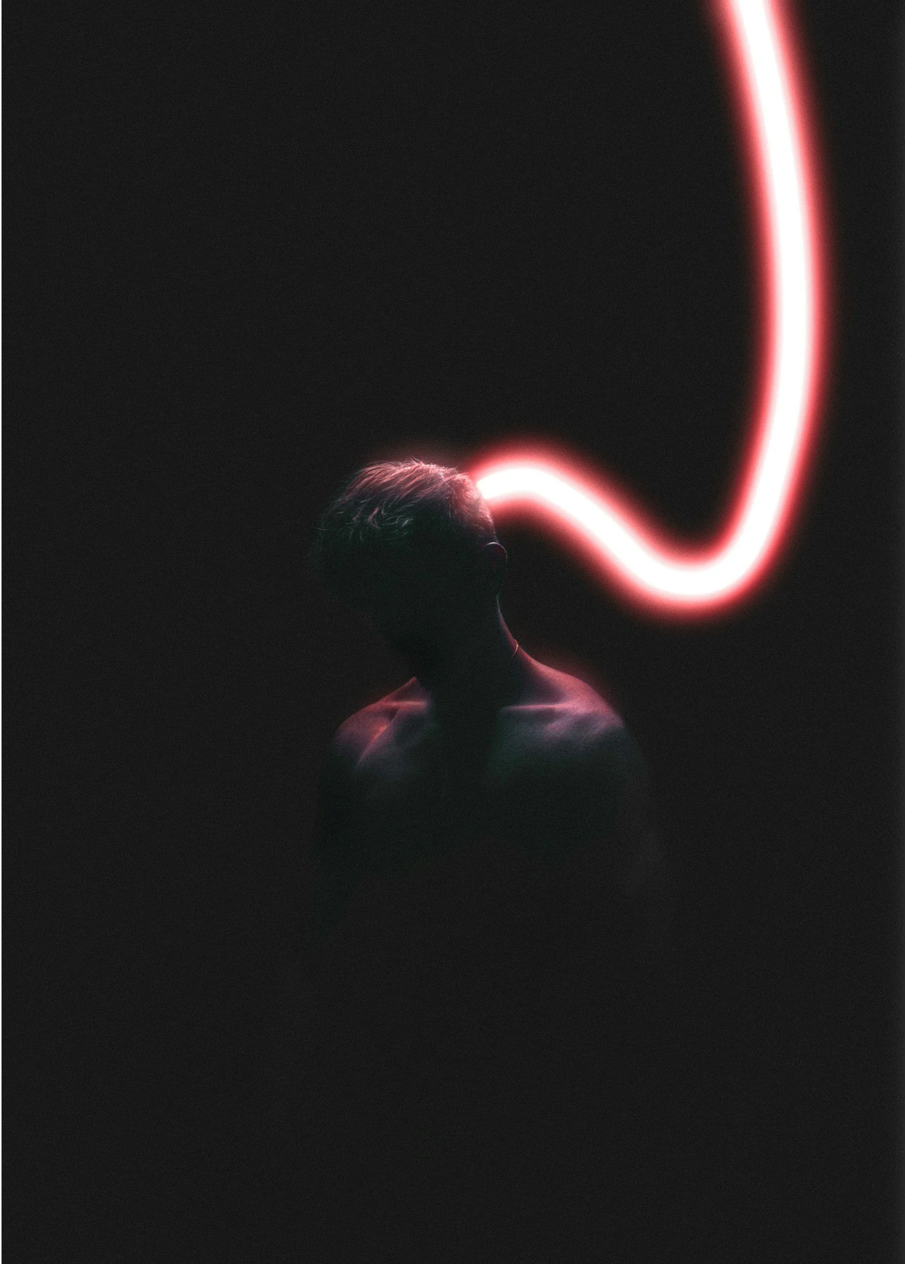 Silhouette of a person with short hair, illuminated by a pink neon light in the shape of a curved line behind them, on a dark background.