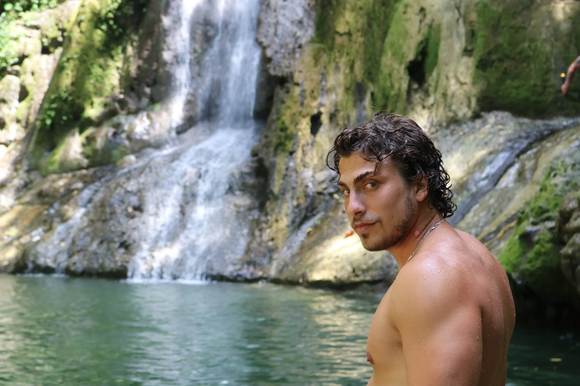 A man with wet dark curly hair and a silver necklace standing in a body of water near a waterfall surrounded by green moss and rocks.
