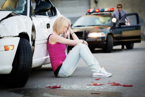 A woman sitting on the ground with a damaged white car beside her, holding her head, after a car accident. A police car is parked in the background with an officer standing nearby.