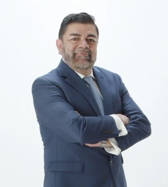 A middle-aged man with dark hair and a beard, smiling, wearing a navy blue suit and tie, crossing his arms against a plain white background.