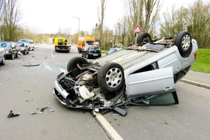 A silver car flipped upside down on the road with extensive front-end damage, surrounded by debris after a crash, with parked cars and trees in the background.