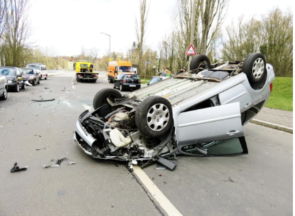 A silver car overturned on its roof in the middle of the road after a crash, with debris scattered around.