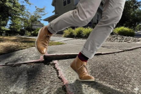 Person walking on a cracked sidewalk wearing tan boots, gray pants, and red socks, with a house and trees in the background.