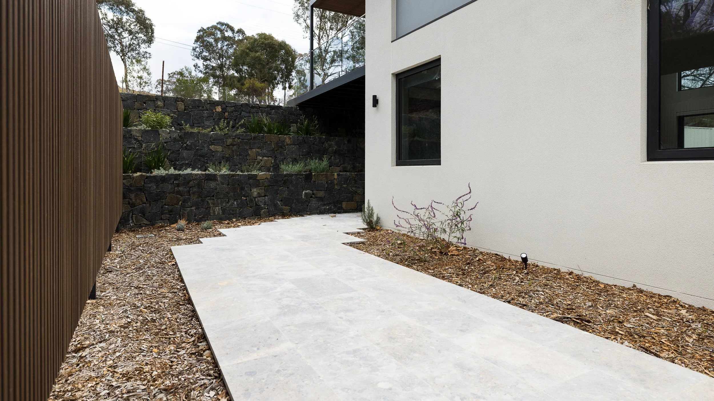 Modern backyard with a concrete pathway, wooden fence on the left, white house wall on the right, and a stone retaining wall with plants in the background.