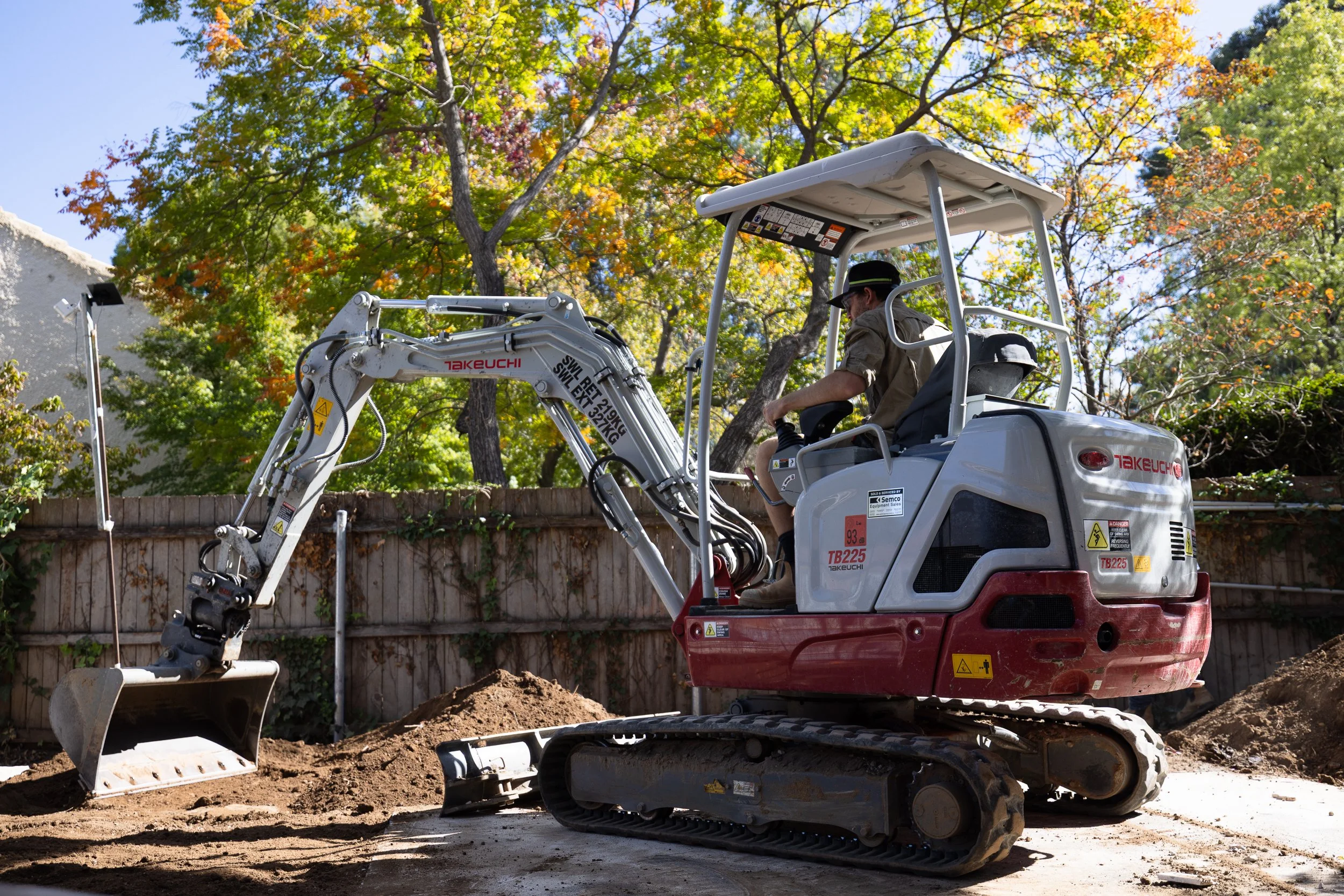 A construction worker operating a mini excavator on a dirt surface behind a wooden fence, with autumn-colored trees in the background.