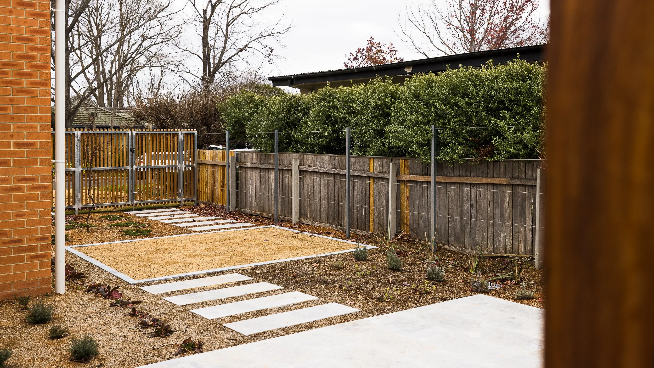 Backyard with a brick wall on the left, gravel and stepping stones walkway, small plants, wooden and metal fencing, green bushes, trees, overcast sky.