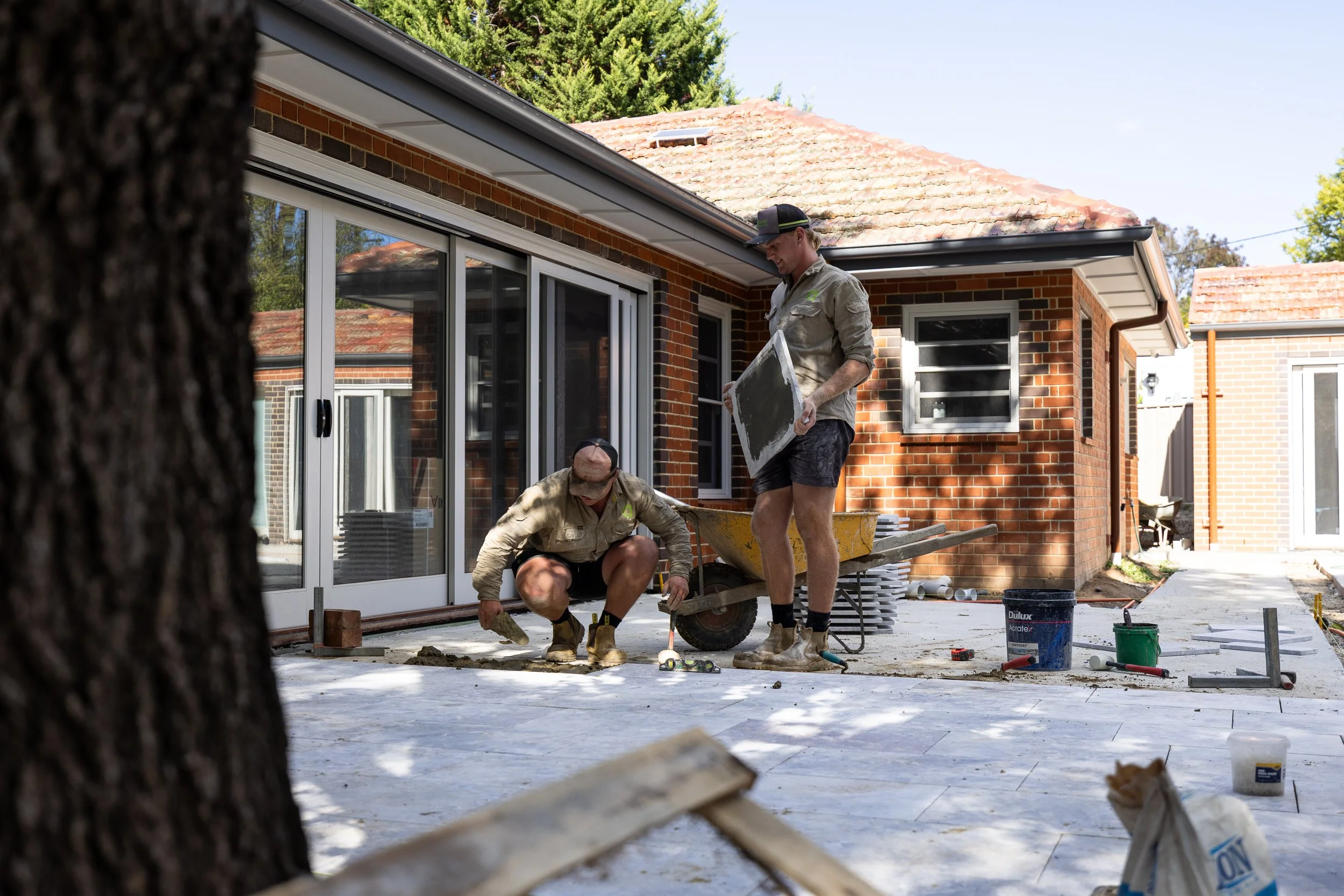 Two men working on laying stone tiles on a patio outside a brick house, with construction tools and materials around them.