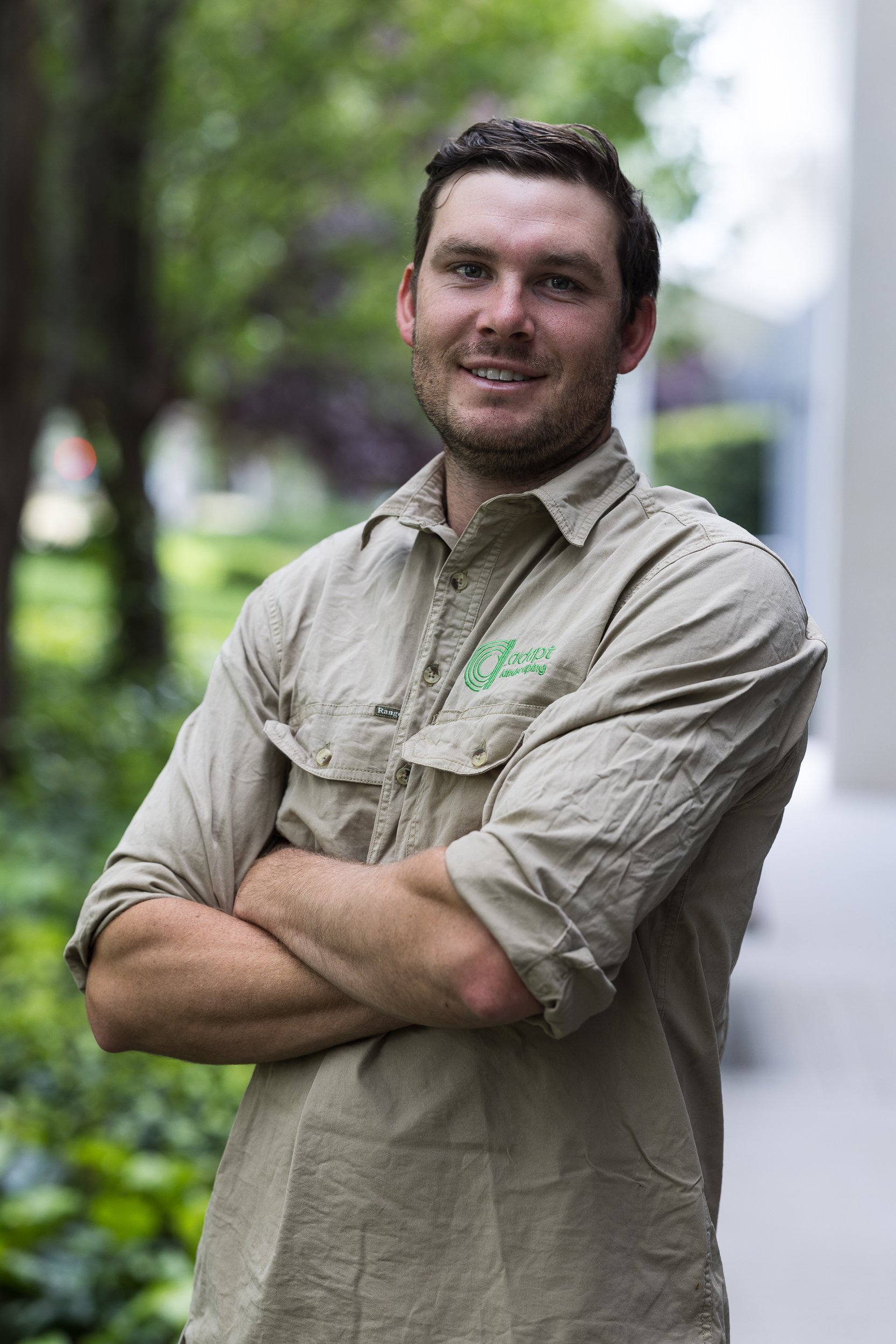 A man with dark hair and a beard smiling with arms crossed, wearing a khaki shirt with green embroidery, outdoors with trees and greenery in the background.
