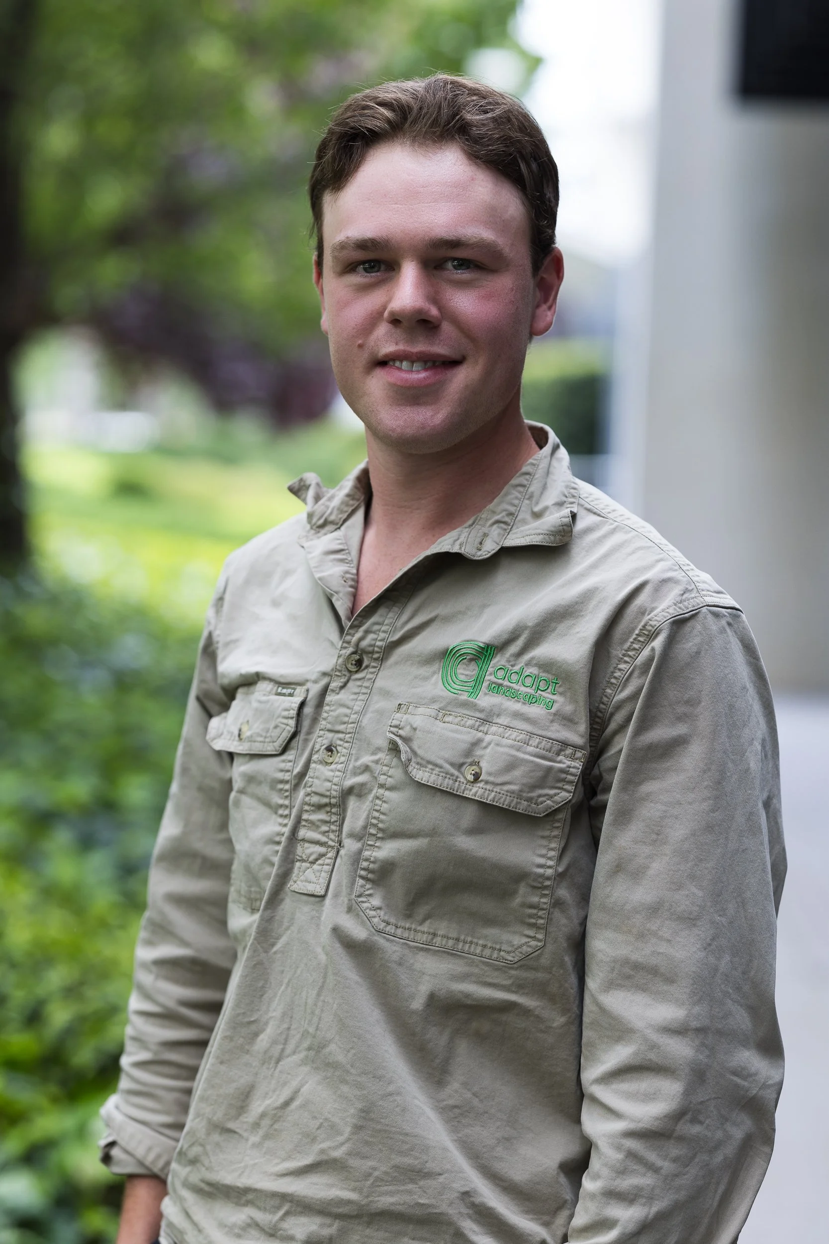 A man wearing a beige shirt with upturned sleeves, standing outdoors with a blurred green landscape in the background.