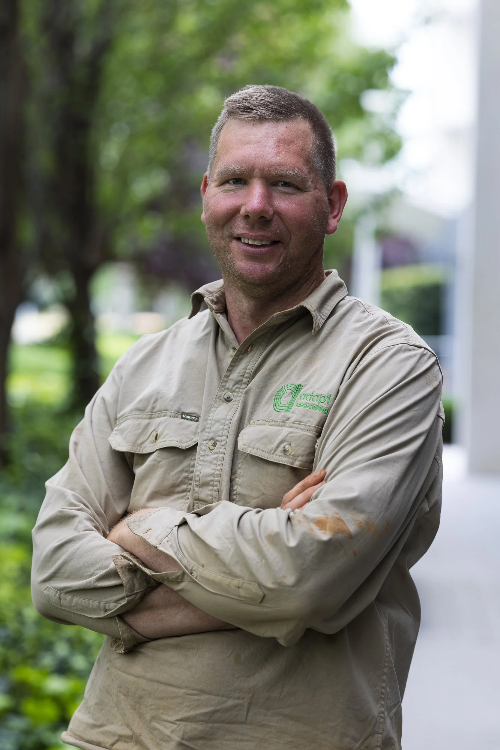 A smiling man with light brown hair wearing a beige work shirt with green embroidery and crossed arms stands outdoors with trees and a house in the background.
