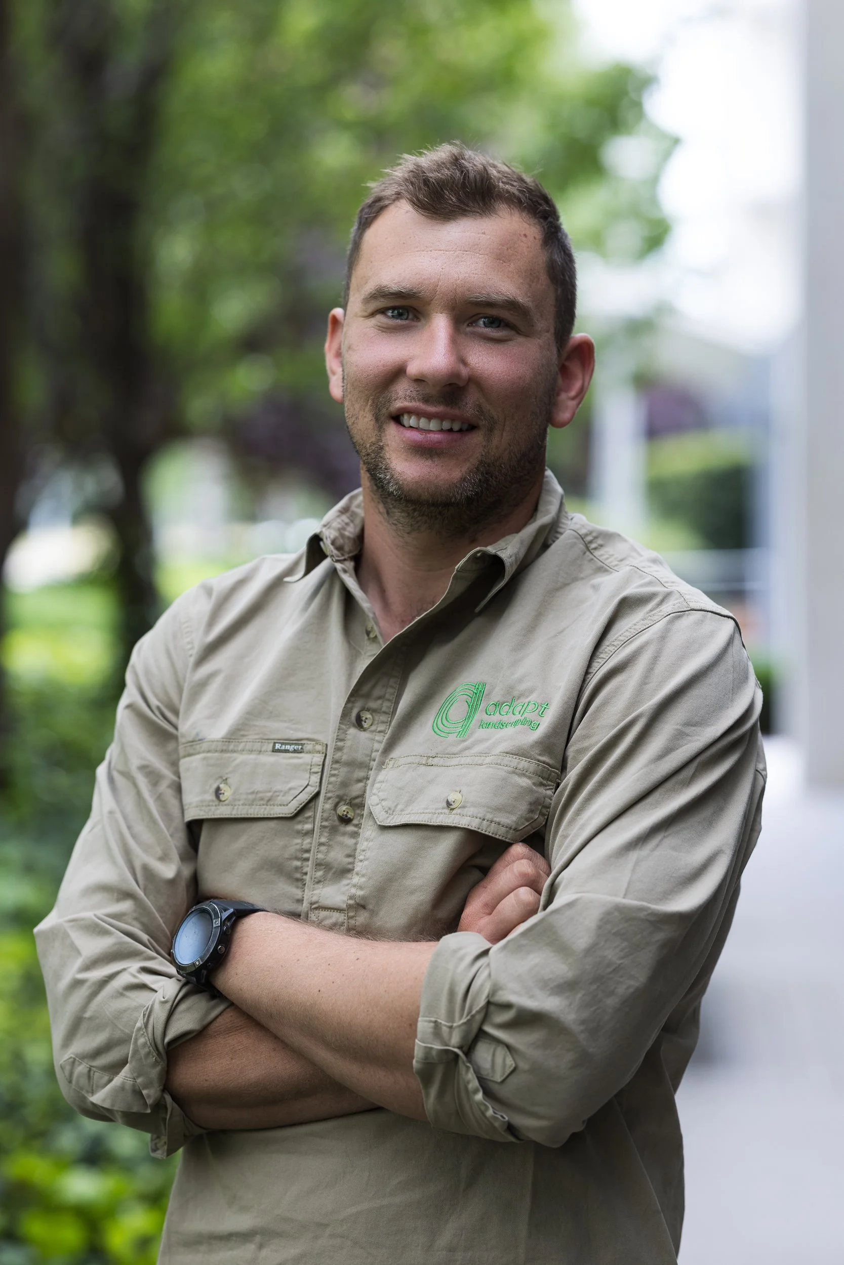 A man with short brown hair, a beard, and blue eyes standing outdoors with trees in the background. He is smiling, wearing a beige button-up shirt with a logo on the chest and a black watch on his left wrist.