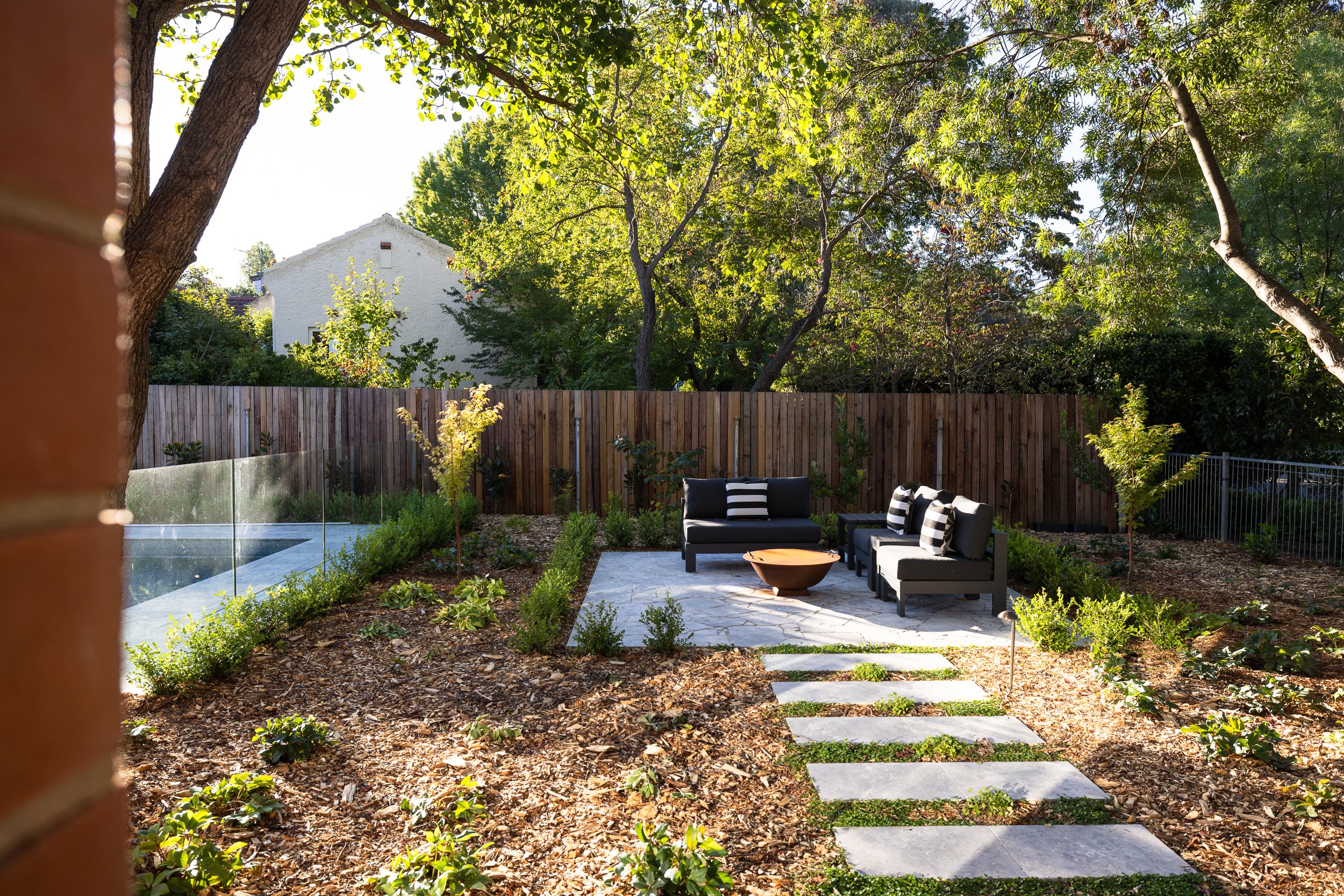 Construction worker operating a mini excavator in a backyard, with green and orange autumn trees in the background.
