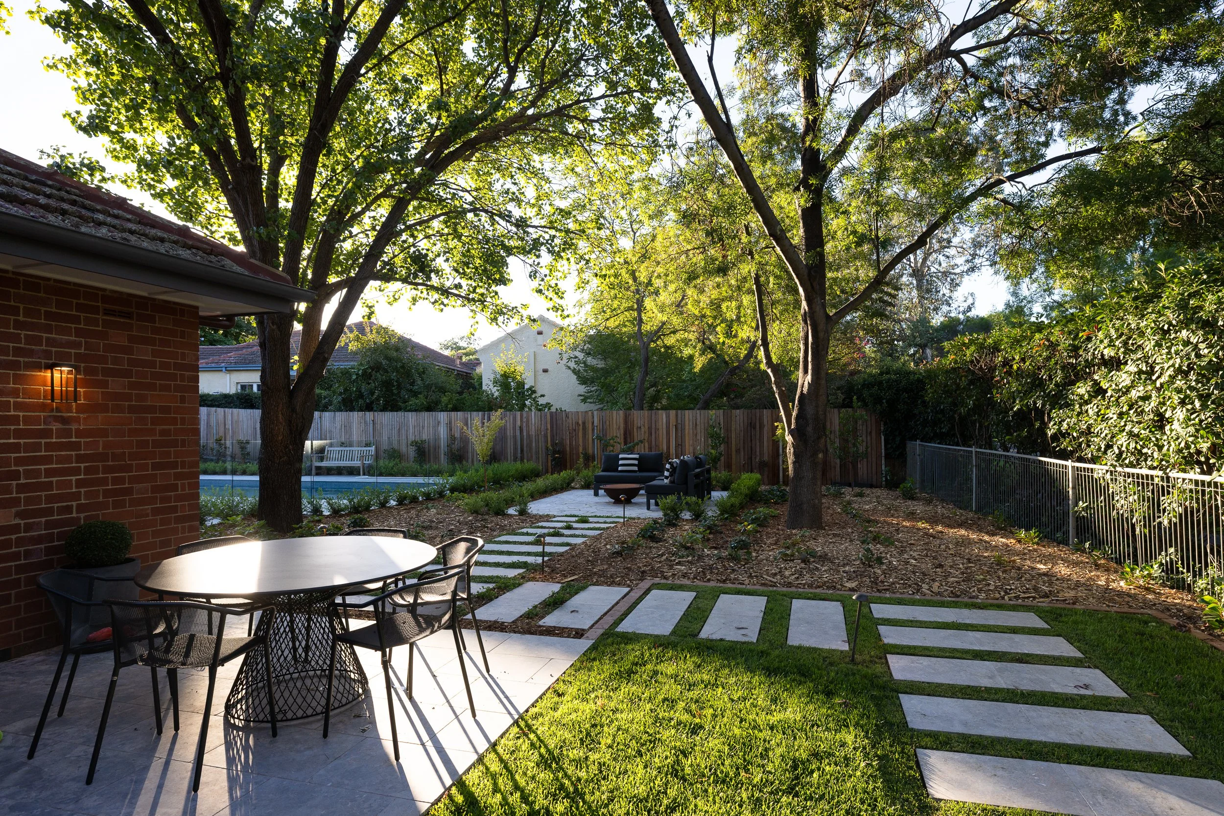 A backyard with a concrete patio, outdoor dining table with chairs, a grassy area, a walkway made of stepping stones, mature trees, a sitting area with outdoor furniture, a fence, and a swimming pool in the background.