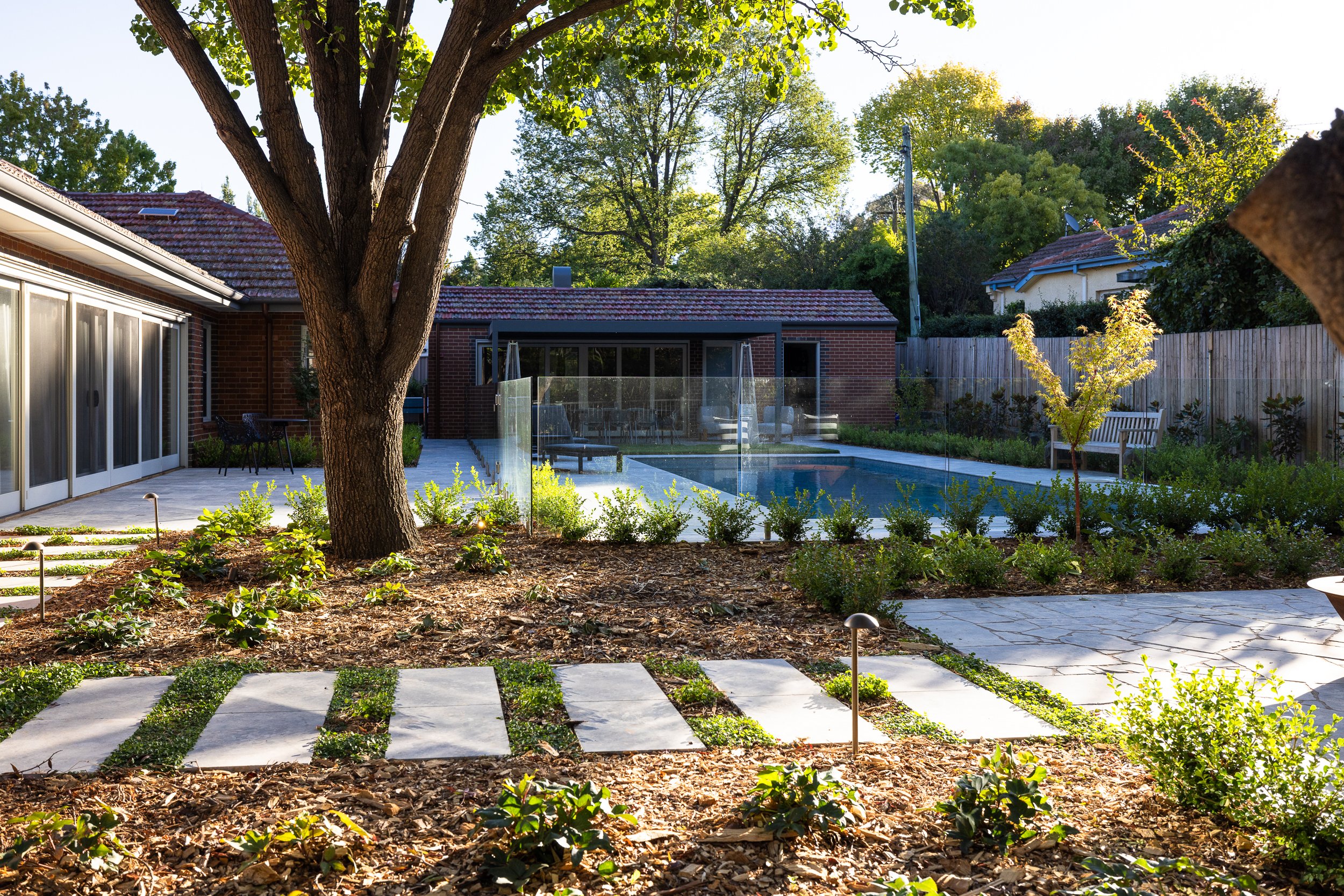 A backyard with a large tree, paved walkway, garden beds, a swimming pool, and a screened patio, surrounded by a wooden fence and neighboring houses.