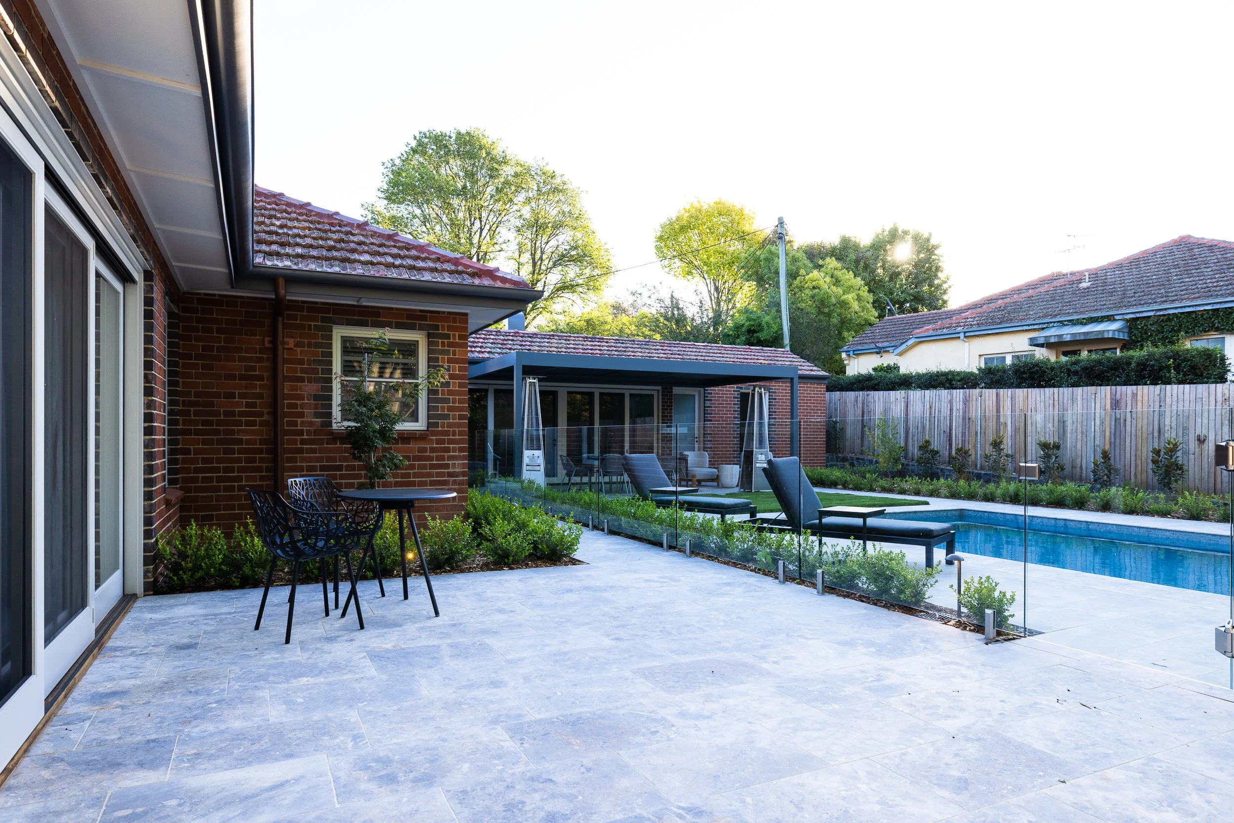 A backyard patio with lounge chairs, a swimming pool, and outdoor furniture, enclosed by a wooden fence, with trees and neighboring houses in the background.