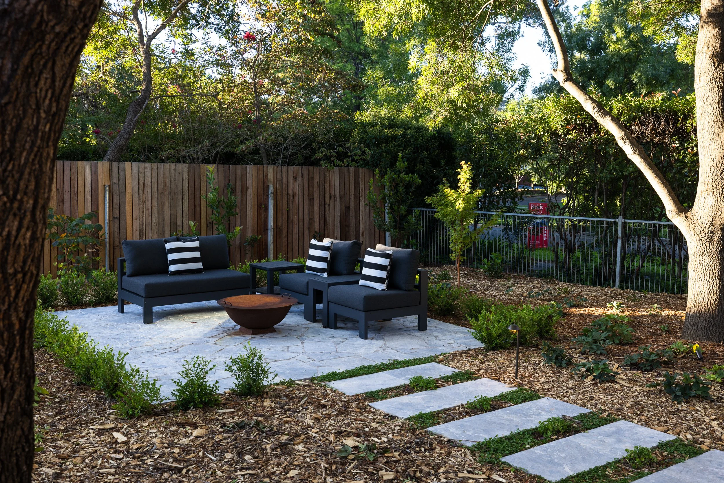 Outdoor patio with black seating and striped pillows, stone pathway, small plants, trees, wooden fence, and garden lighting in a backyard
