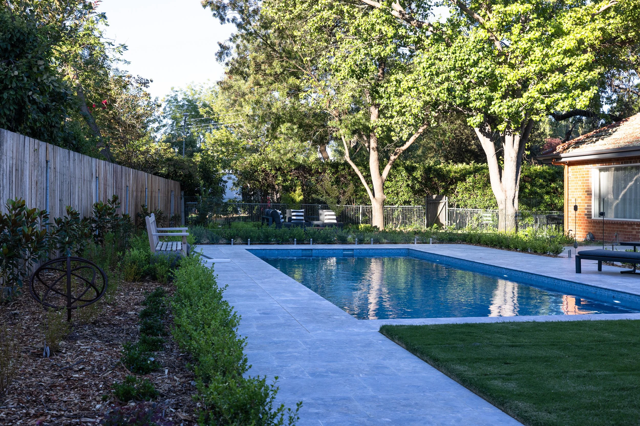 Residential backyard with a rectangular swimming pool, surrounded by a stone patio, green grass lawn, trees, and outdoor seating.