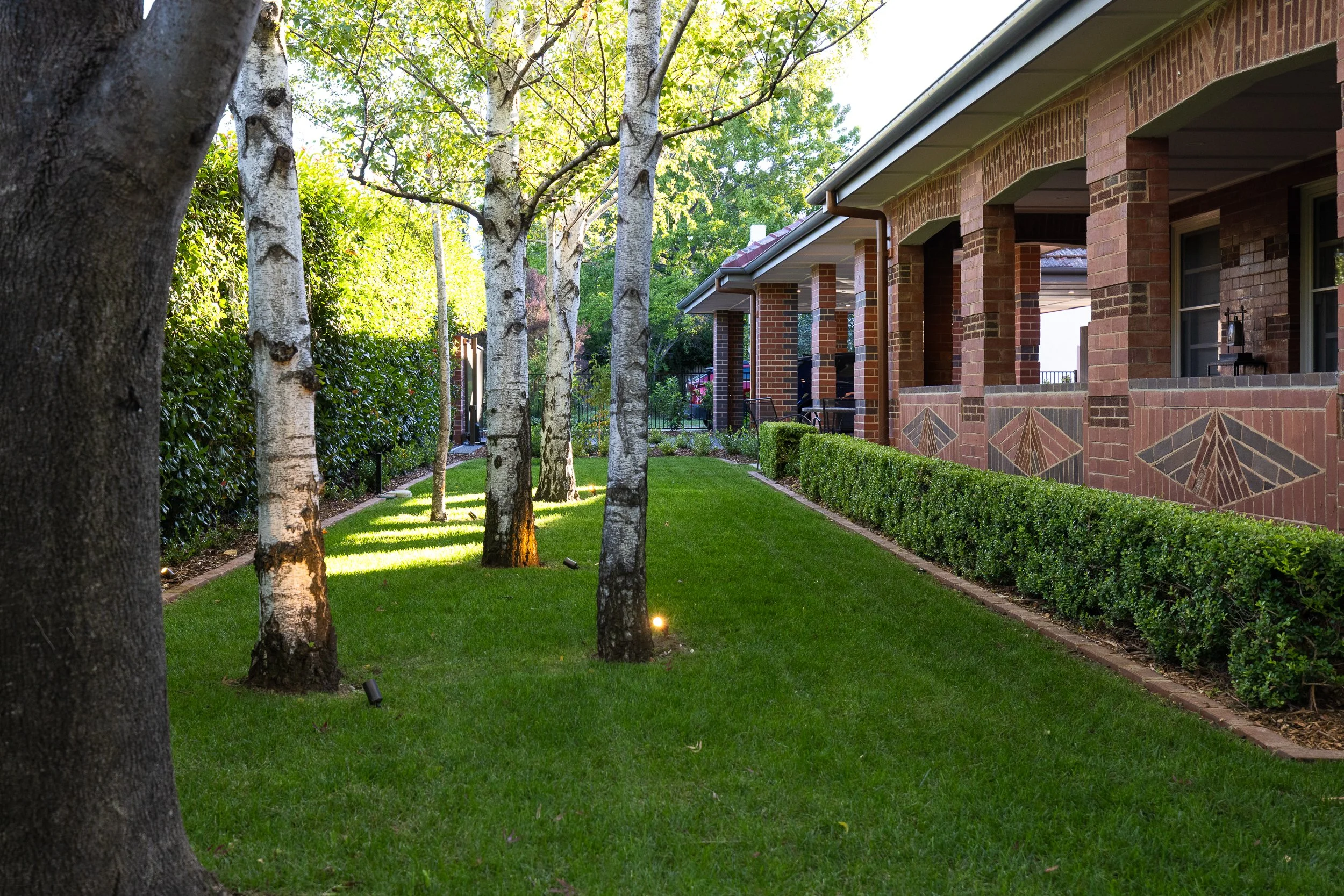 Well-maintained backyard with green grass, tall trees, trimmed shrubs, and a brick house patio with columns.