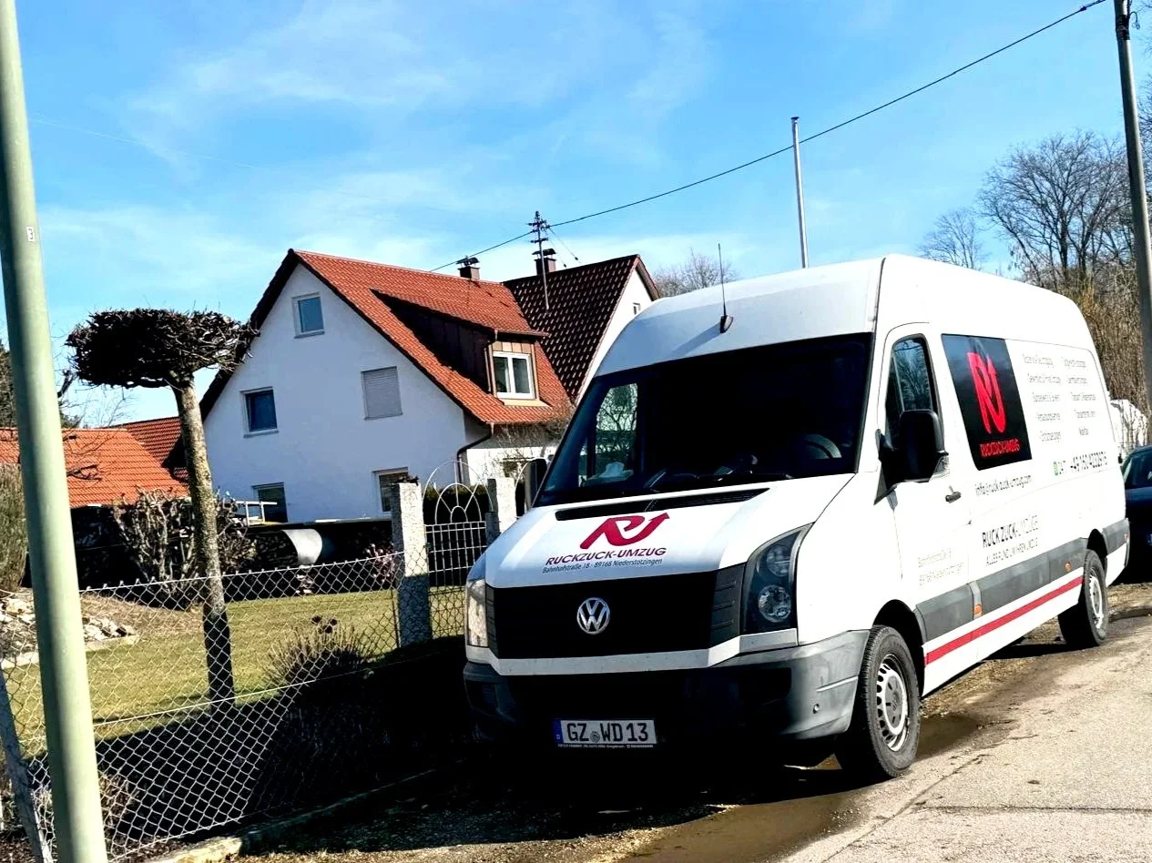 Weiße Umzugsfirma-Transporter mit Logo und Kontaktinformationen parkt neben einem Zaun vor einem Haus mit rotem Dach, auf einer Straße bei sonnigem Wetter.