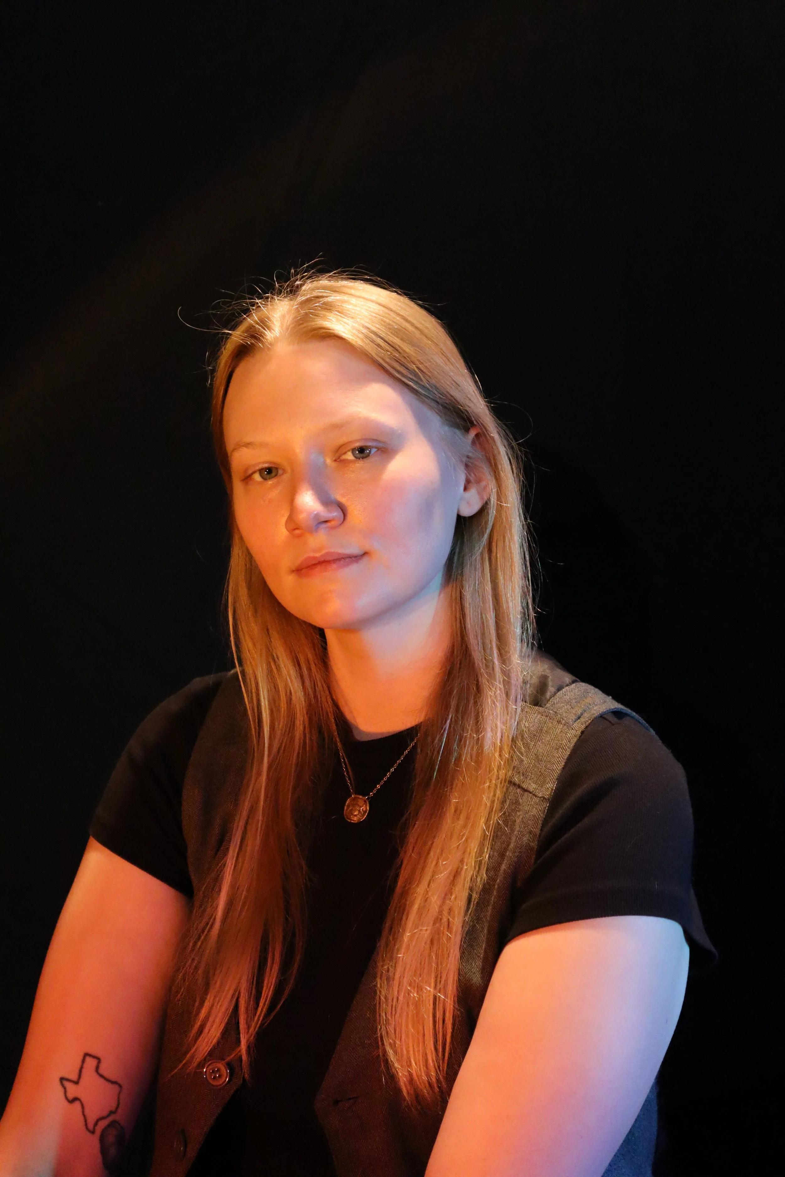 A young woman with long red hair, wearing a black t-shirt and a pendant necklace, poses against a dark background.