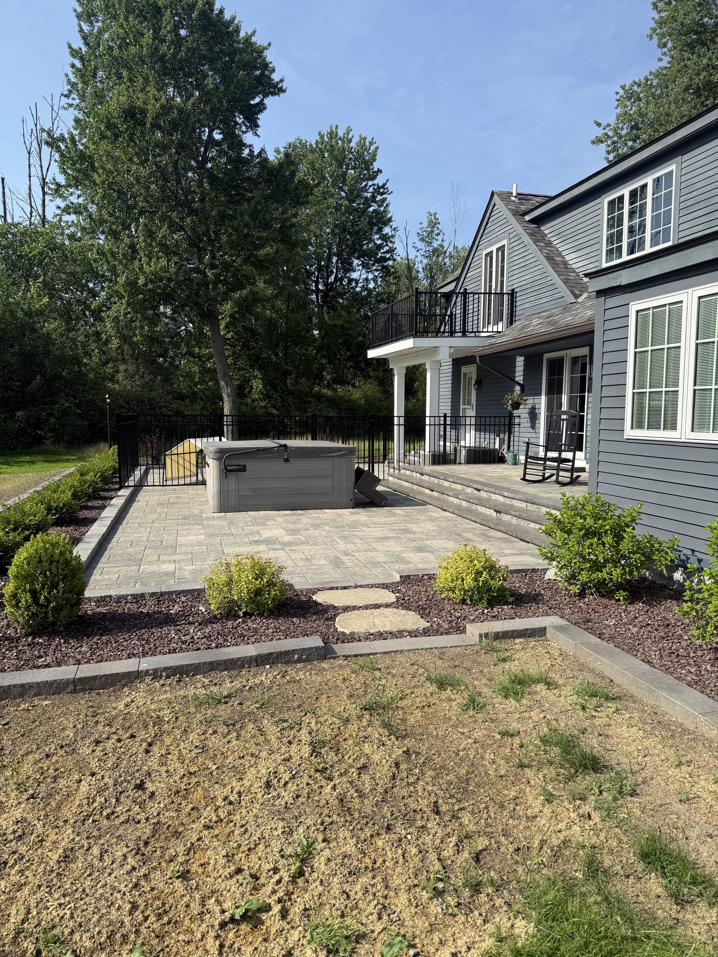 Backyard with a paved patio, hot tub, a few succulent bushes, and a house with grey siding and multiple windows, under a clear blue sky.