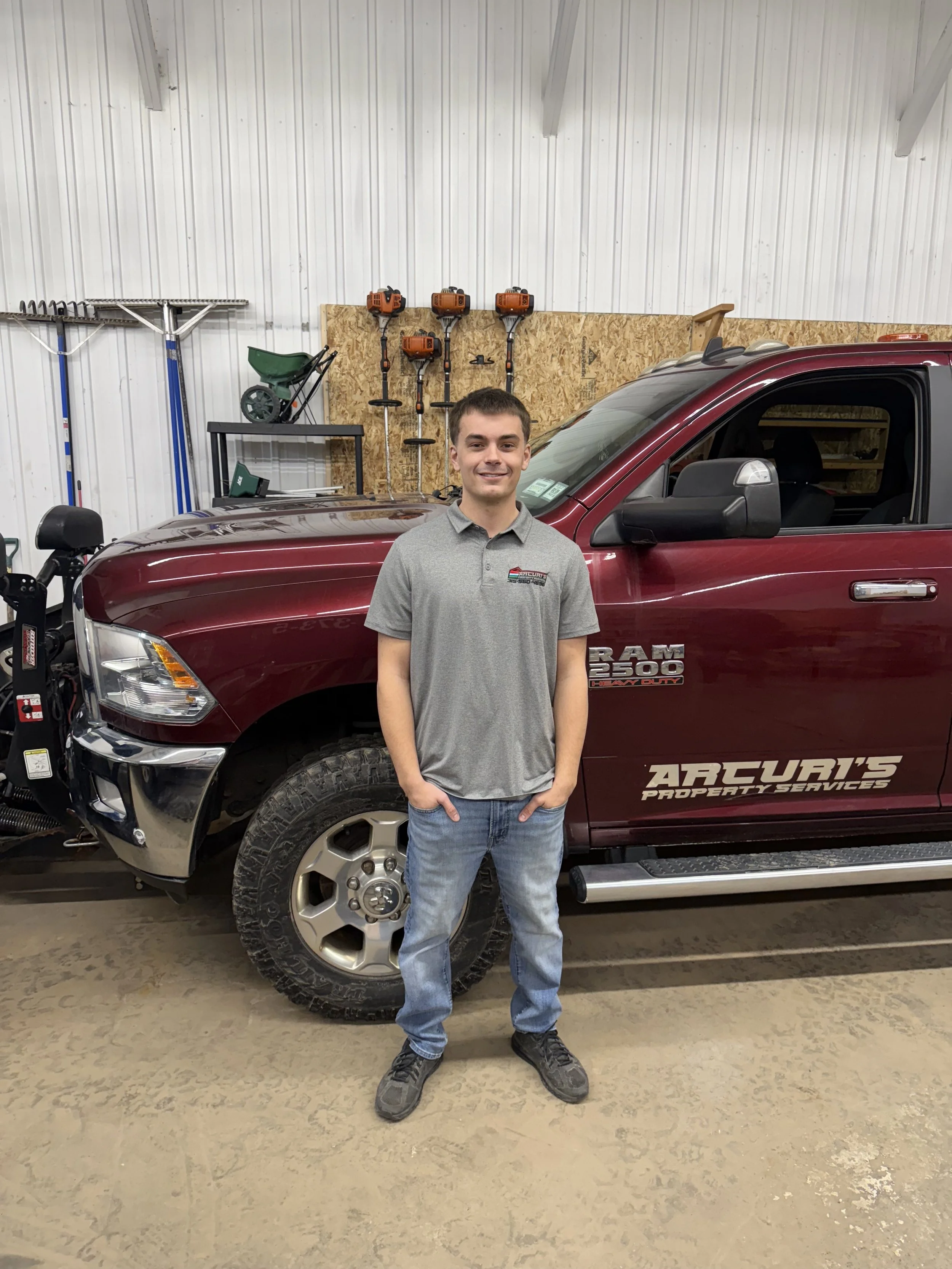A young man in a gray polo shirt and jeans standing in front of a maroon RAM 2500 truck inside a garage or workshop. The background includes tools and equipment on the wall, such as trimmers and a wheelbarrow.