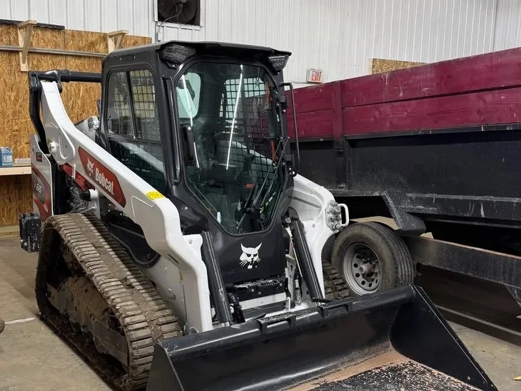 A Bobcat compact track loader parked indoors next to a black and red utility trailer.