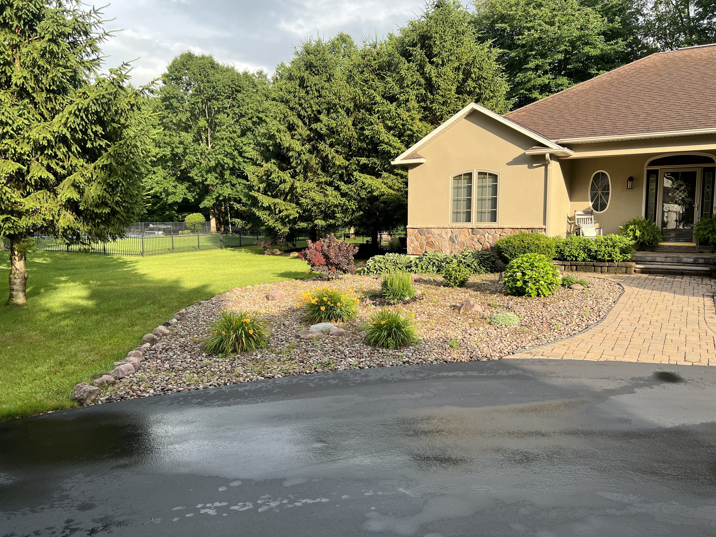Front yard with well-maintained lawn, decorative flower bed, trees, and a house with a stone and stucco exterior, brick driveway, and porch area.