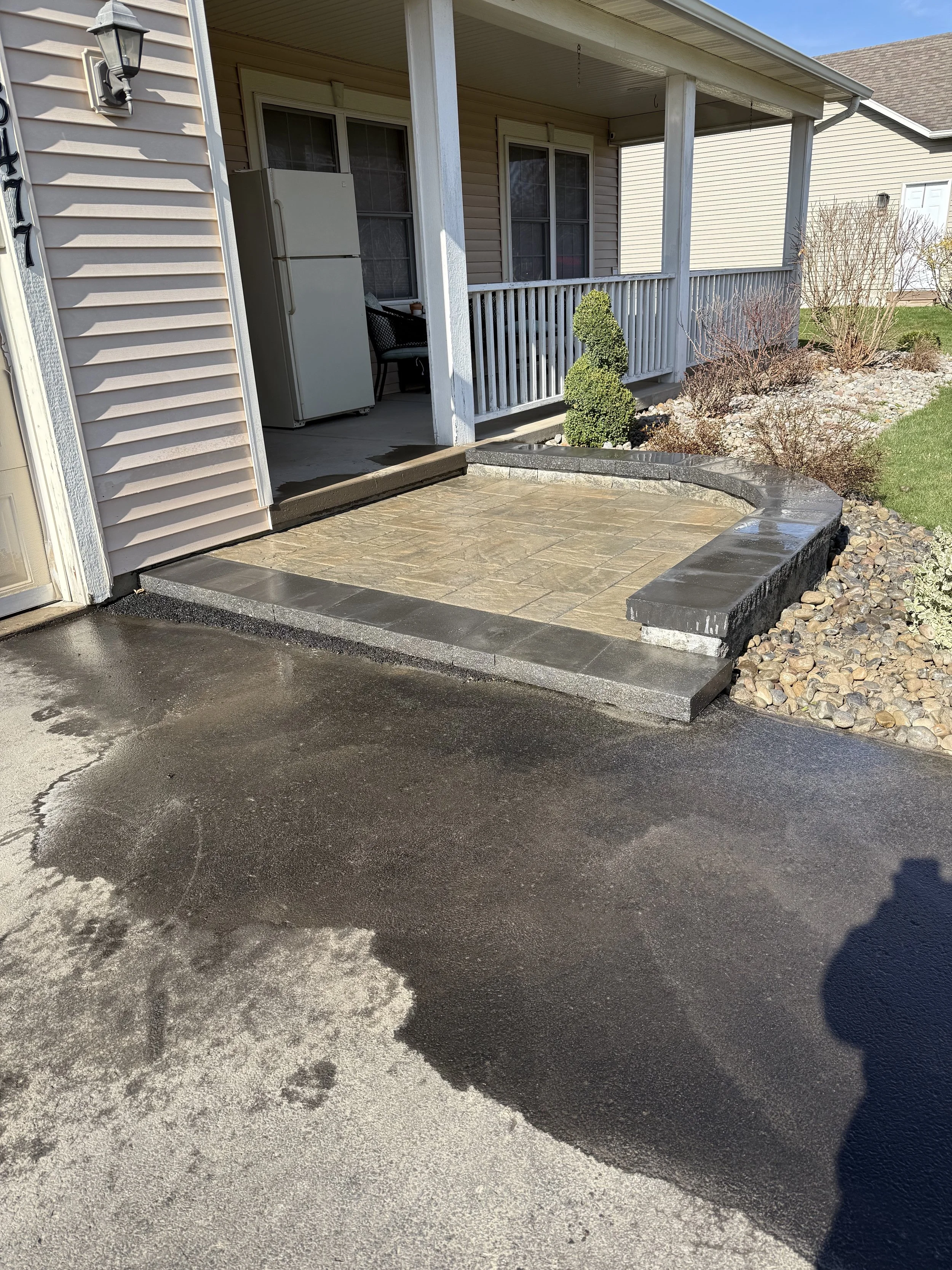 Front porch with newly paved entryway, two steps leading to a small balcony, a refrigerator on the porch, and landscaped bushes and rocks.