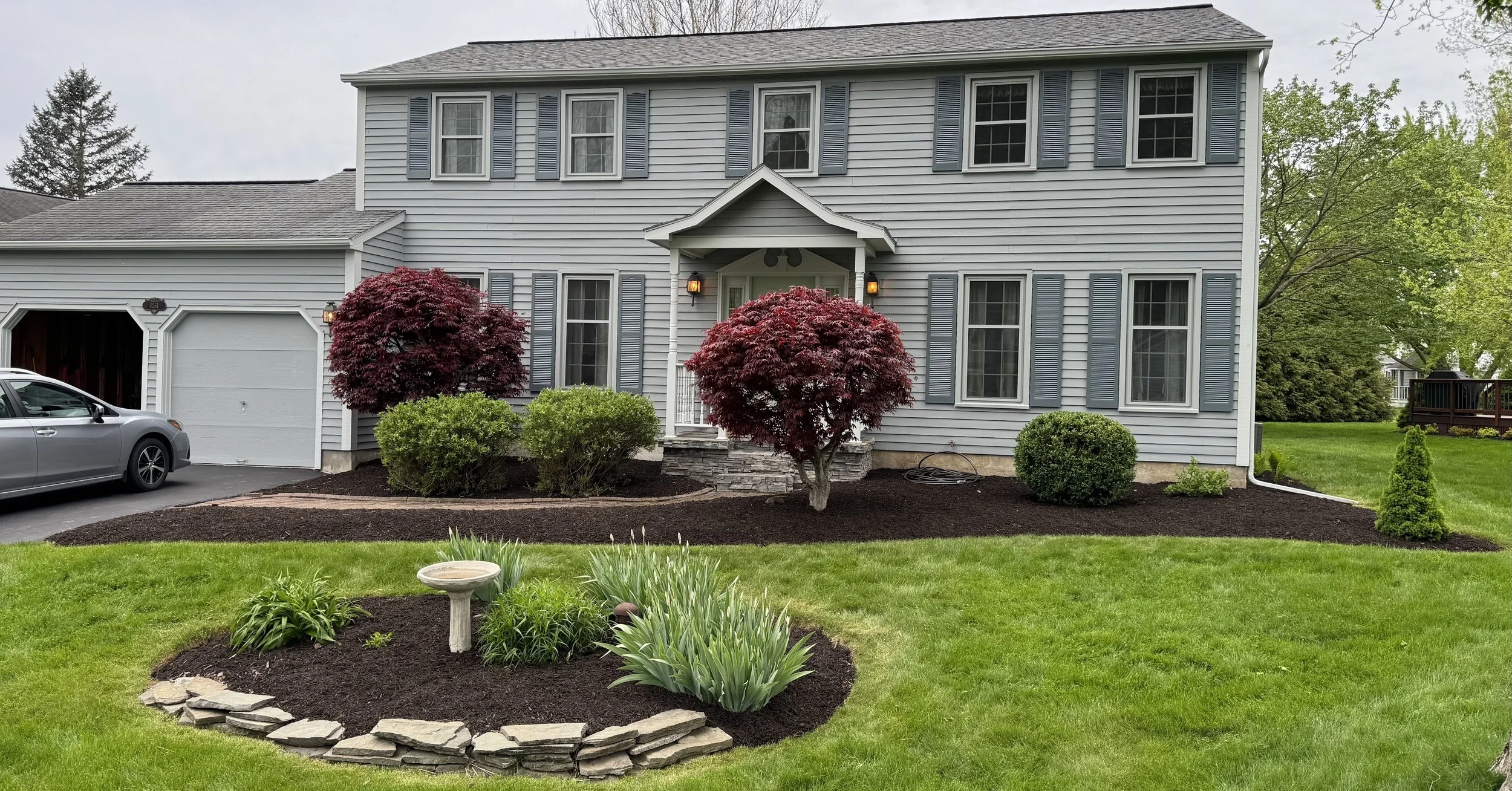 Front yard of a two-story house with gray siding, blue shutters, and a detached garage. The yard features a neatly landscaped garden with bushes, two prominent red-leafed trees, and a small stone-bordered flower bed with a birdbath. A silver car is p
