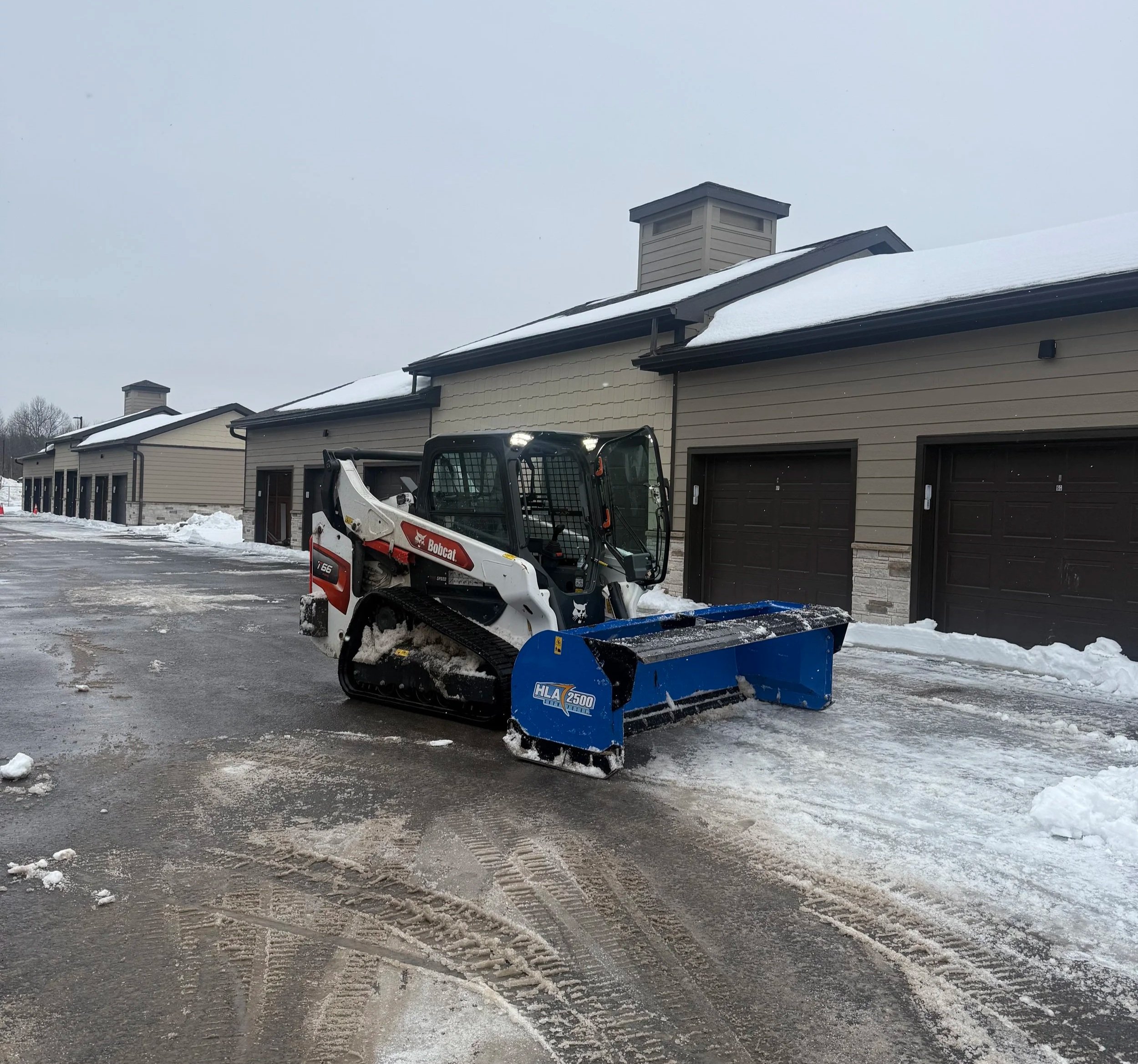 A bobcat snow blower attachment clears snow from a paved driveway in a residential area with multiple garage units and snow on the roofs.