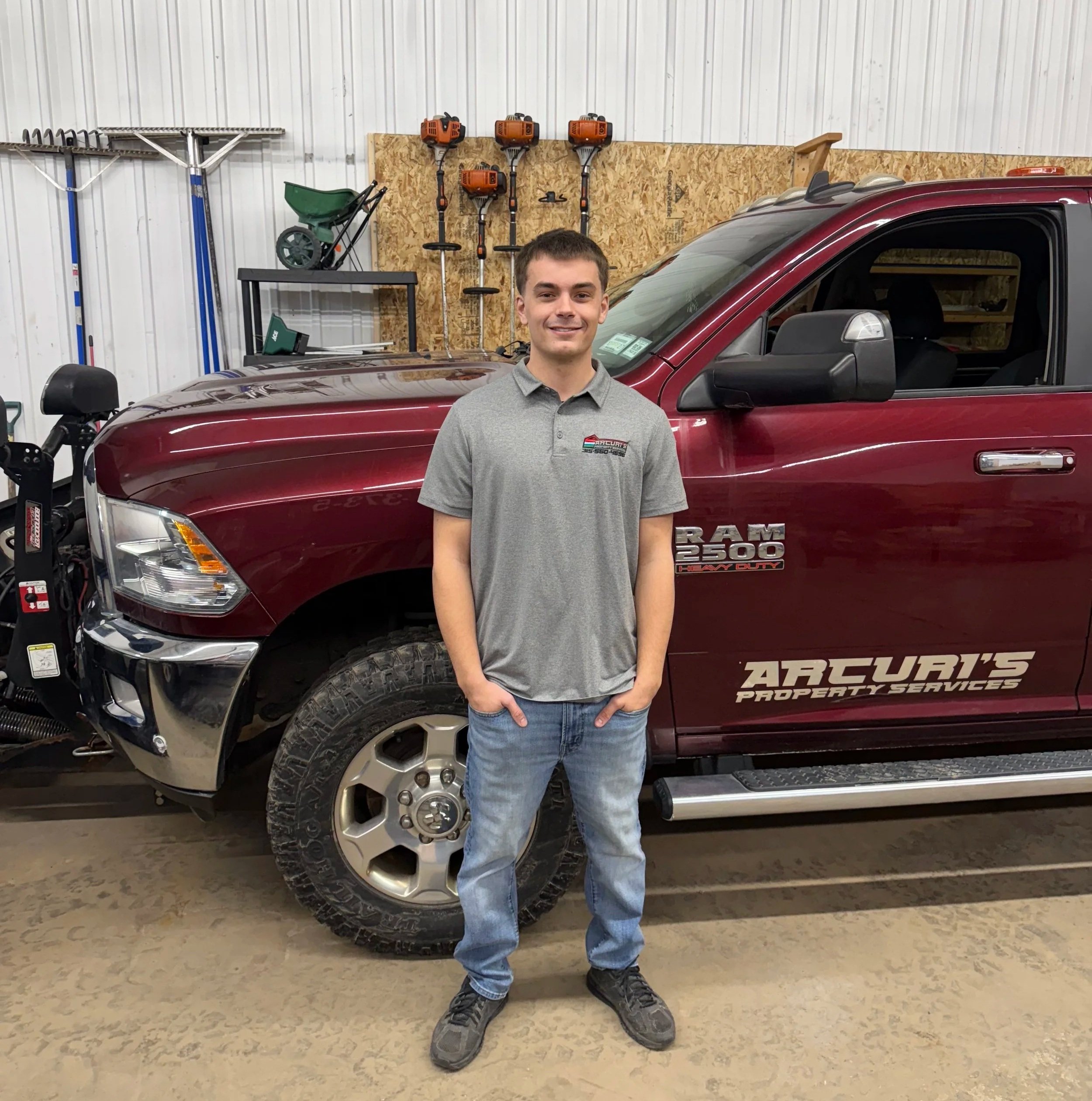 A young man standing in front of a maroon RAM 2500 truck inside a garage or workshop, with various tools and equipment in the background.