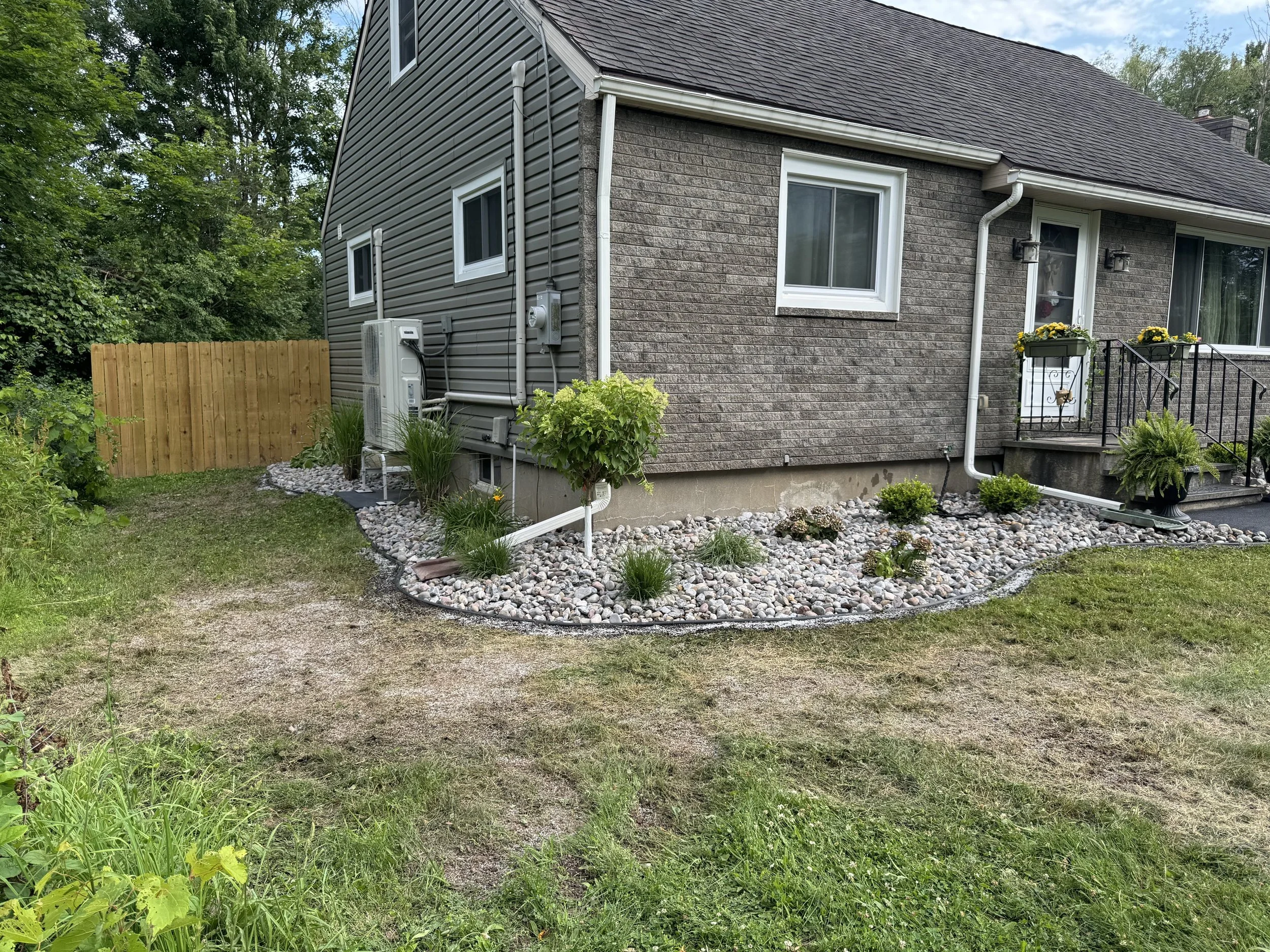 The side yard of a house with a well-maintained stone garden bed, a short set of stairs with black railings leading to a back door, and a wooden privacy fence. The house features brick and grey siding, with drainage pipes and utility boxes visible.