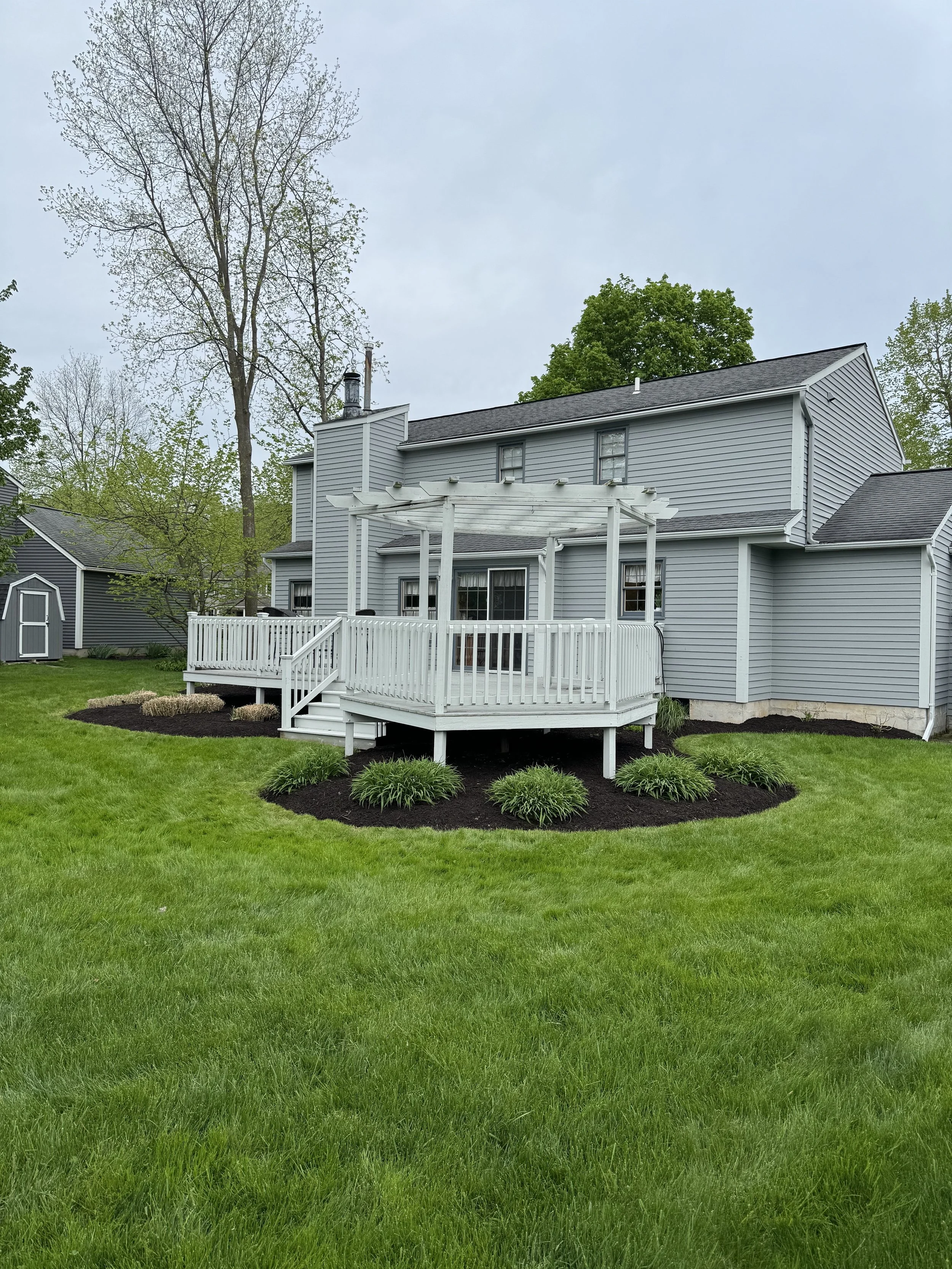 Backyard of a blue house with a white deck and railing, surrounded by green grass and landscaped flower beds with bushes, and tall trees in the background.
