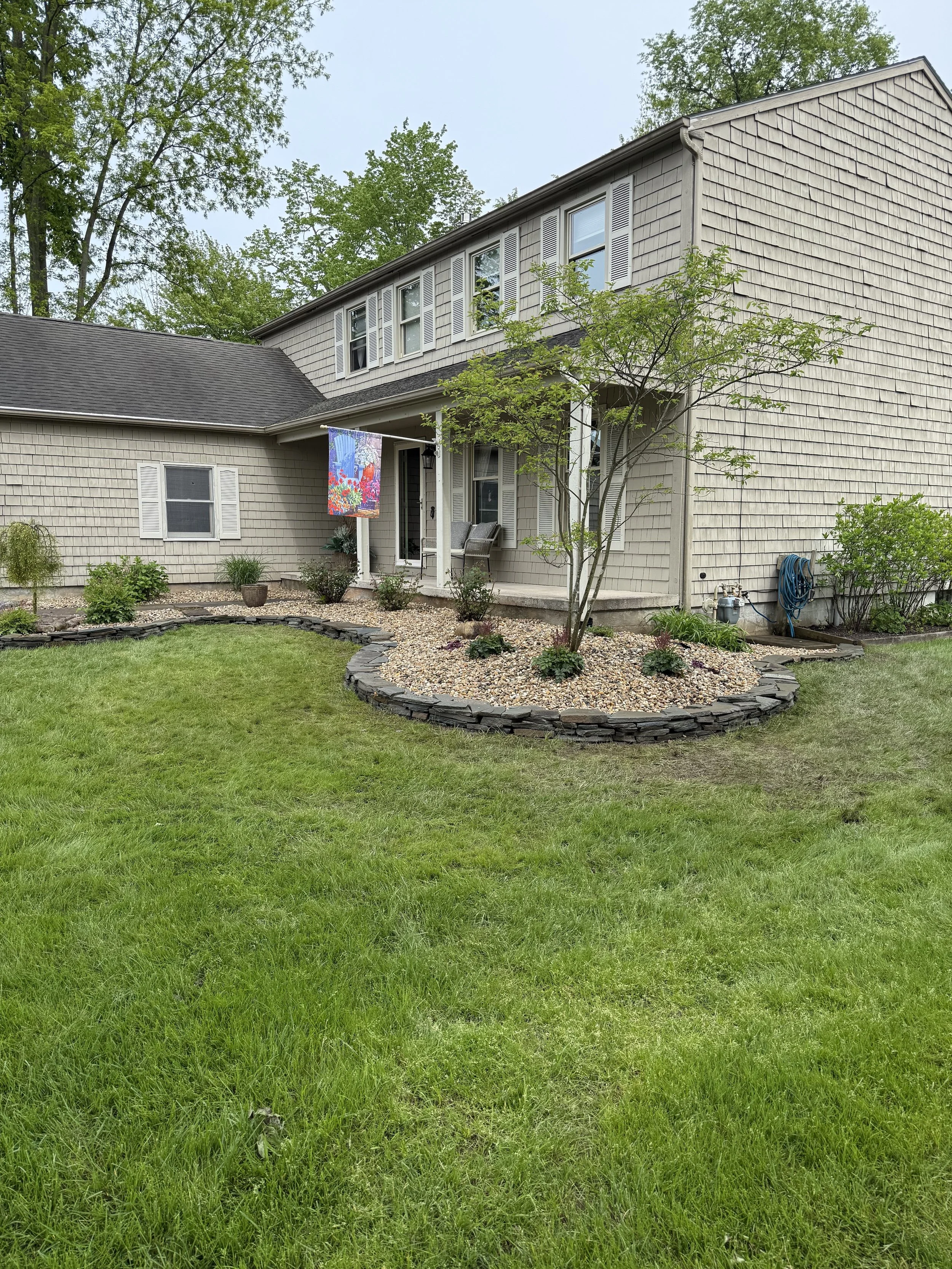 The image shows the backyard of a house with a well-maintained lawn, a small tree, garden beds with shrubs and plants, bordered with stacked stone edging, and a two-story house with beige siding and white shutters. There is a colorful decorative hang