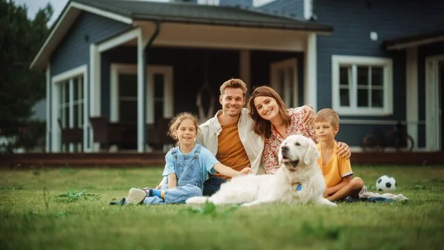 A family of four, including a father, mother, and two children, sitting on the grass with their golden retriever dog in front of a house. There is also a soccer ball nearby.