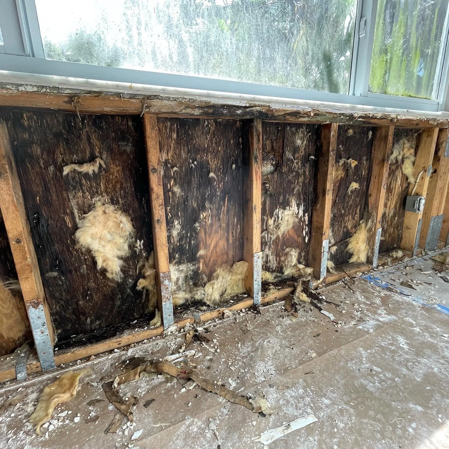 Interior of a damaged wall with exposed wooden studs and blackened wood, likely from fire damage. Yellow insulation material is hanging out and scattered on the floor, which is covered in debris and dust. A window is visible at the top.