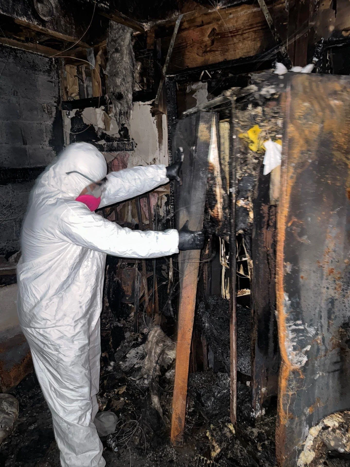 Person in a white protective suit and pink mask using a tool to examine the interior of a burned, charred room.