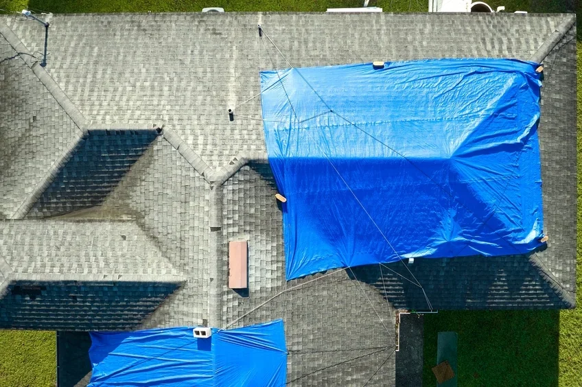 aerial view of a house with a gray shingled roof, and two blue tarps secured with ropes, covering parts of the roof and yard.