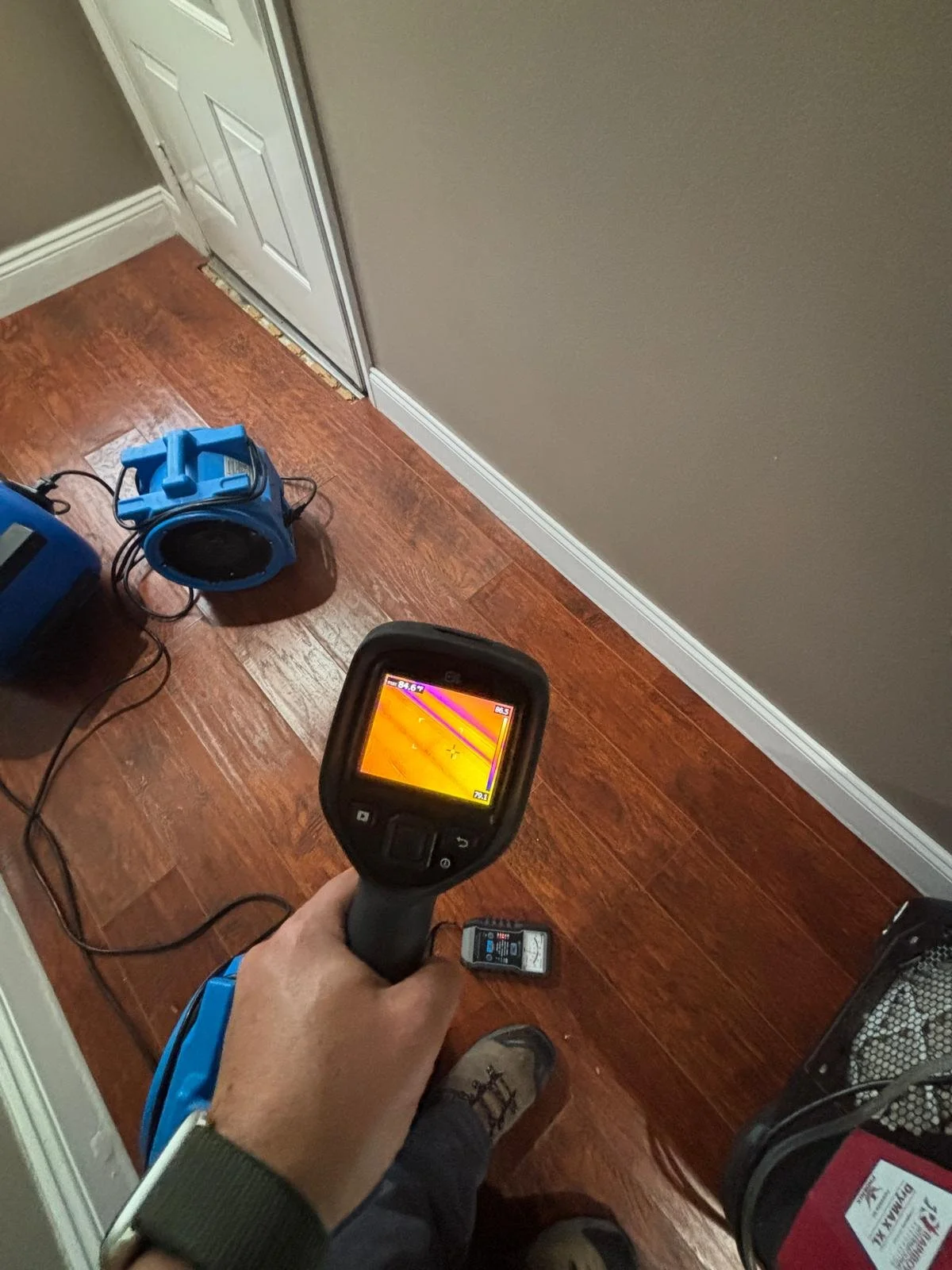 A person using a thermal imaging device to scan a room, showing a color thermal image on the screen, with various equipment and a small device on the wooden floor.