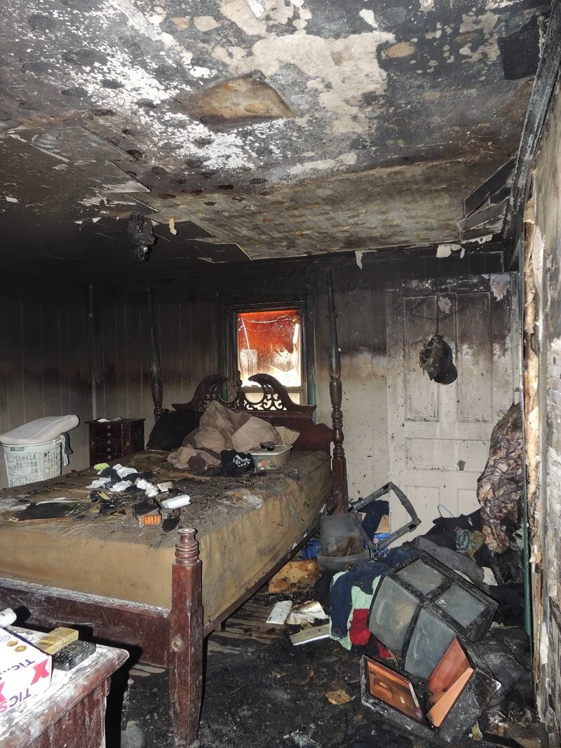 A bedroom with extensive fire damage, including a burned ceiling, a sooty bed, and debris on the floor.